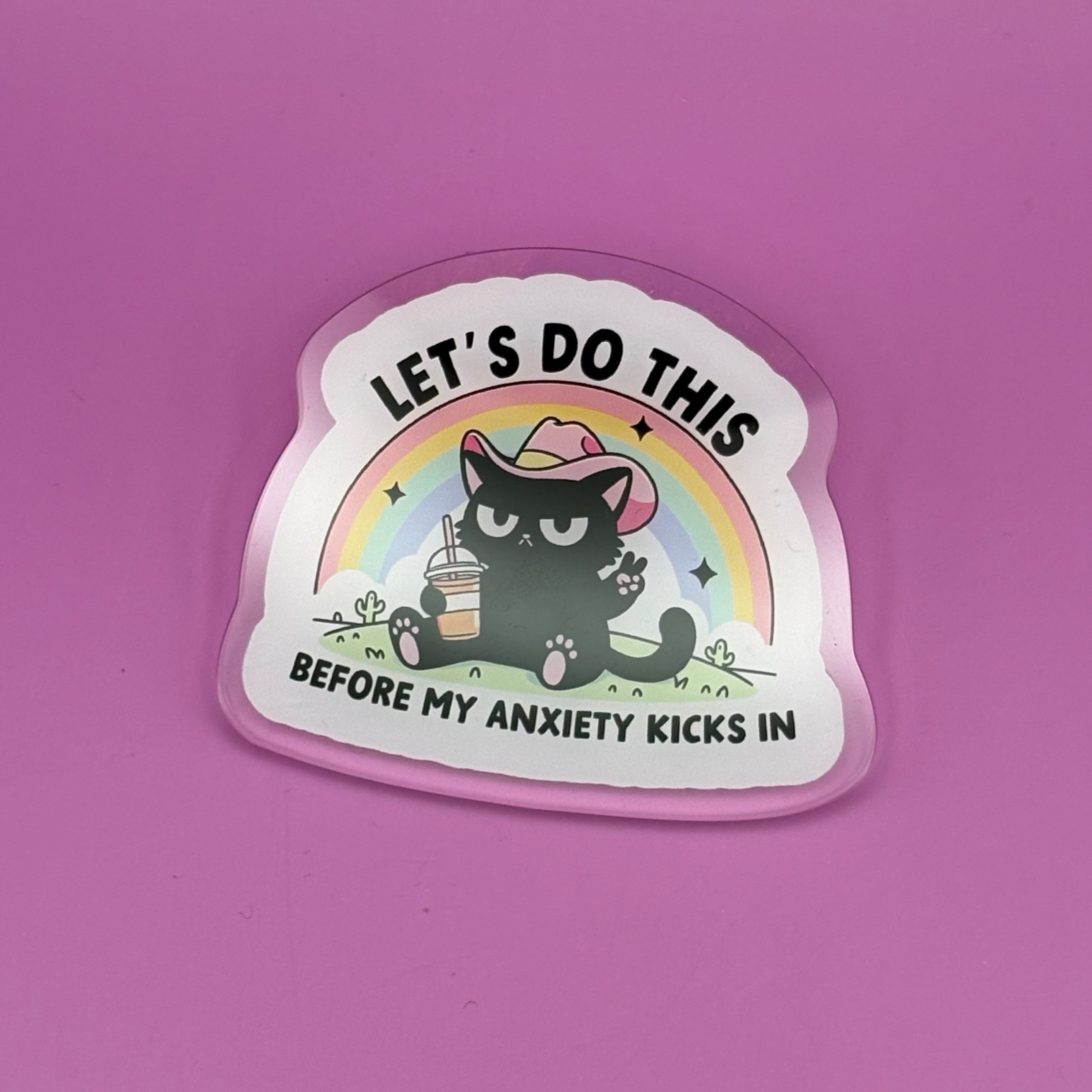 A die-cut sticker featuring a black cartoon cat holding a pink drink in front of a rainbow, with the text 'LET'S DO THIS' and 'BEFORE MY ANXIETY KICKS IN', placed on a solid pink background.