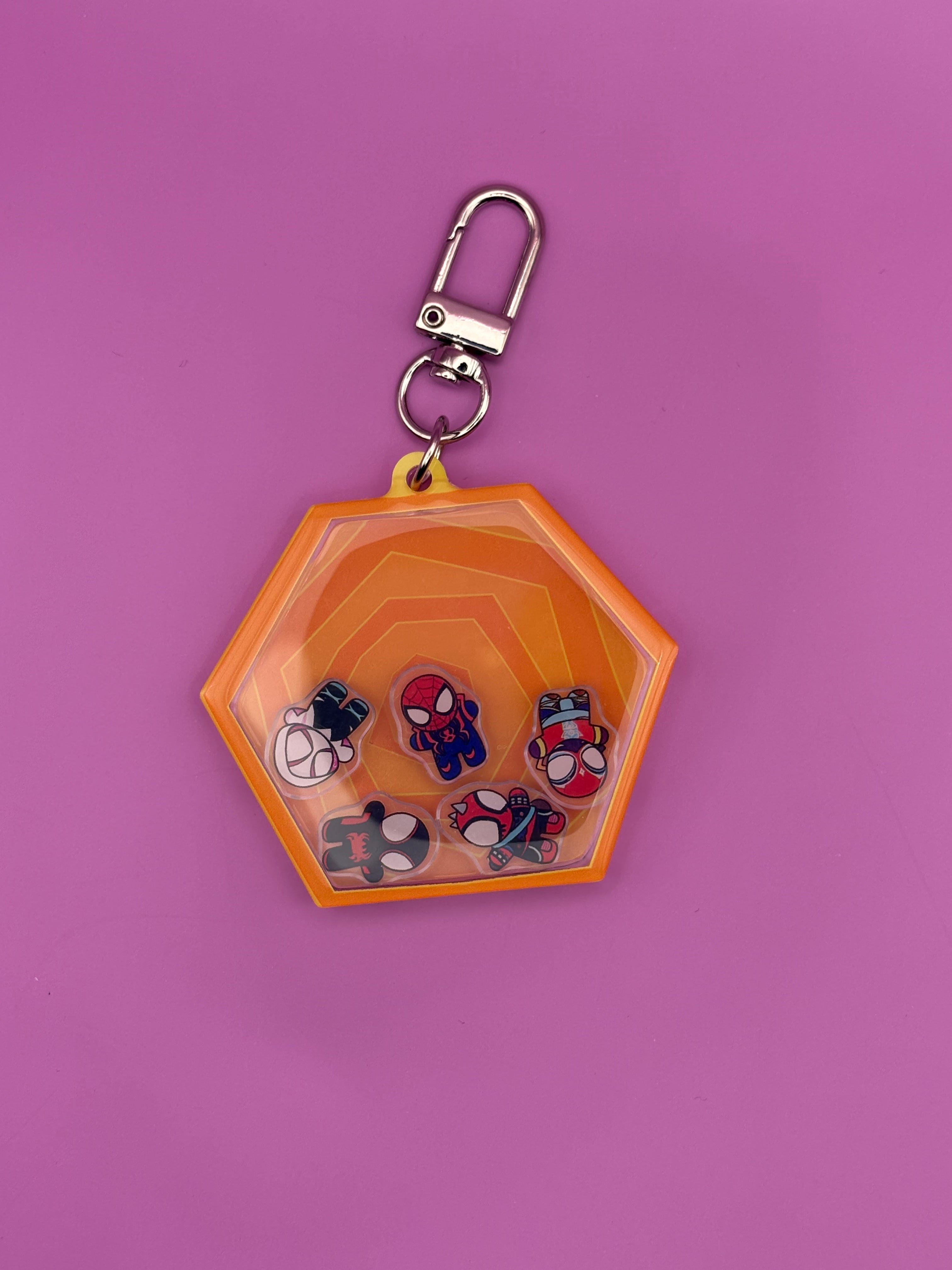 A hexagonal clear plastic keychain holder with a bright orange border, featuring small Spider-Man-themed miniatures inside, attached to a silver metal clasp, set against a solid pink background.