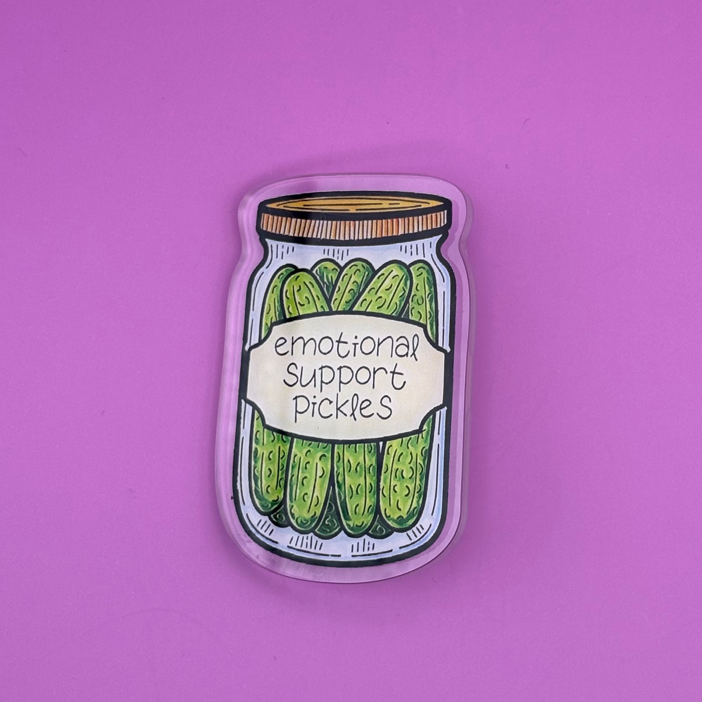 A small enamel pin shaped like a jar of pickles, featuring illustrated green cucumbers and the text 'emotional support pickles,' placed on a solid pink background.