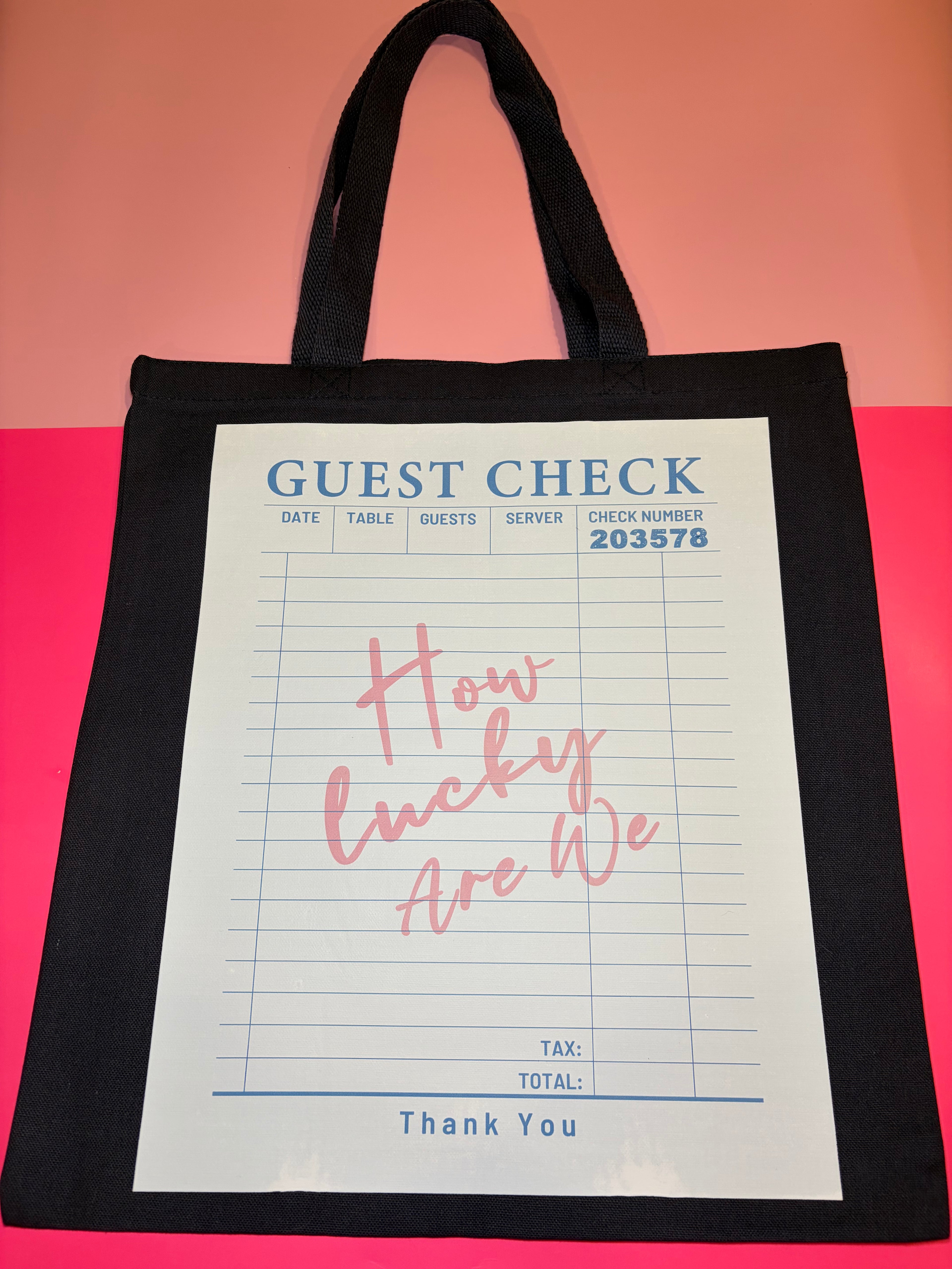 Black tote bag with a printed guest check design with pink "How lucky are we" text