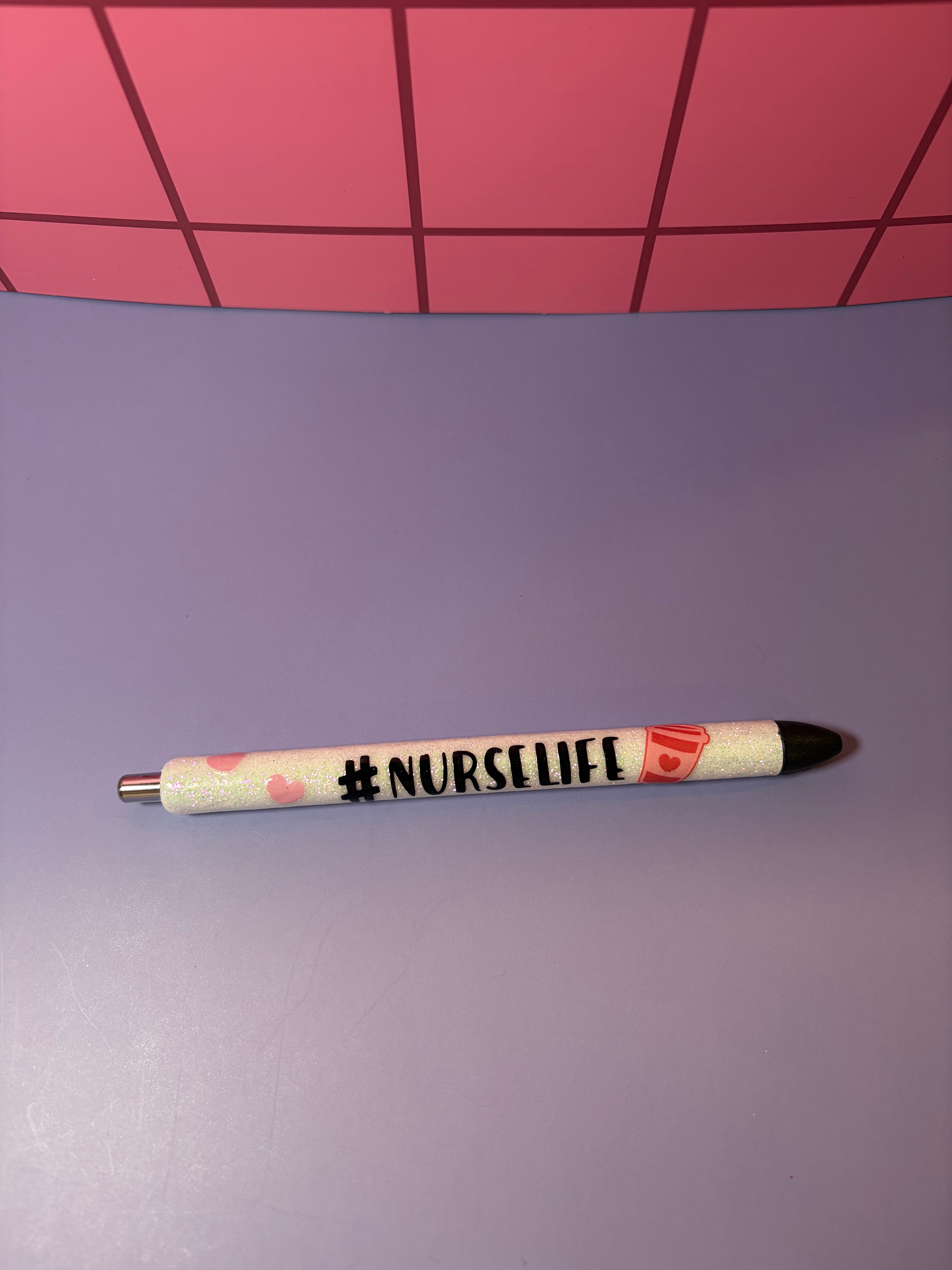 White glitter pen with '#NURSELIFE' text on a purple surface with pink grid background