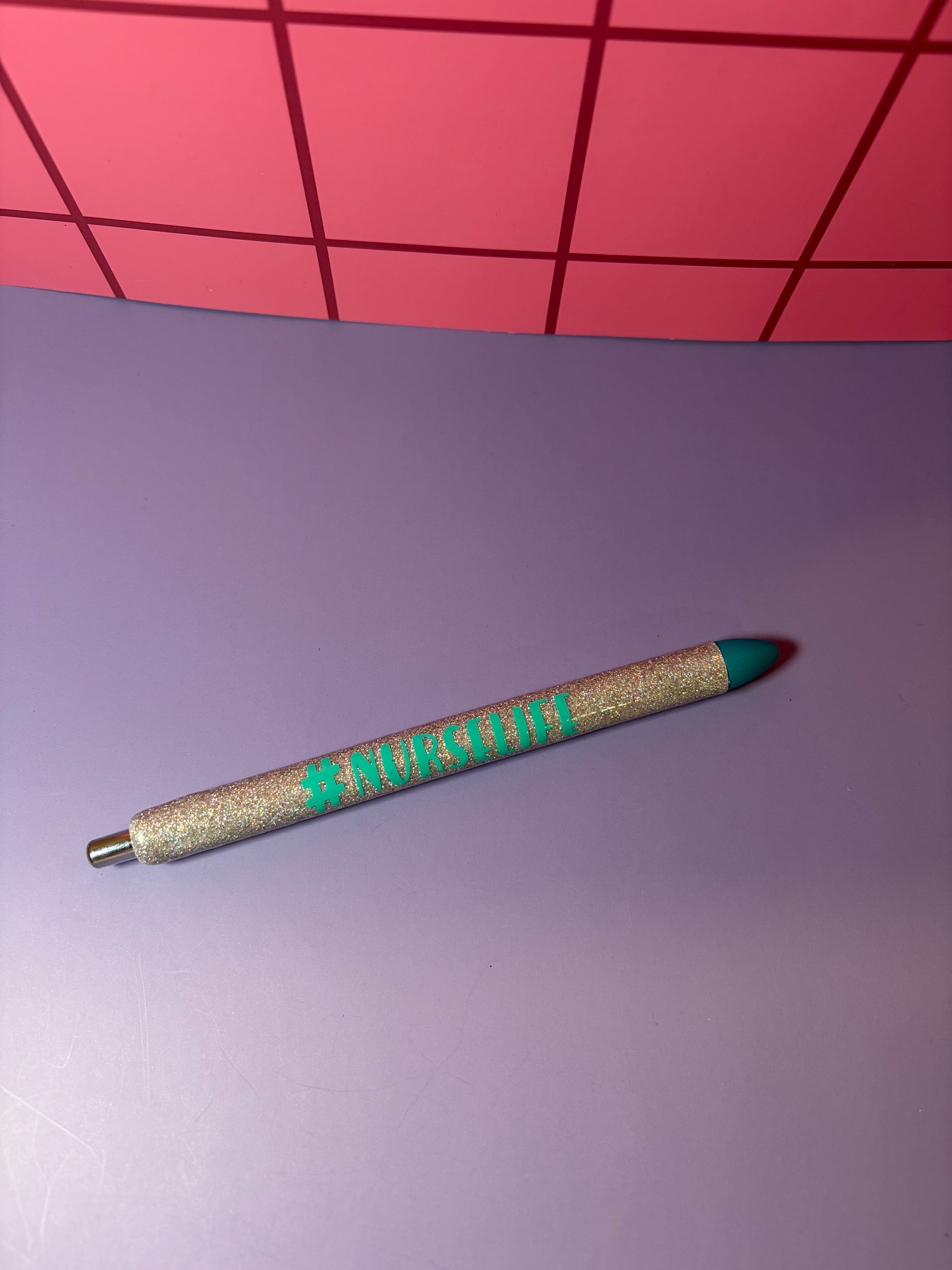 Glittery silver pen with '#NURSELIFE' blue text on a purple surface with pink grid background