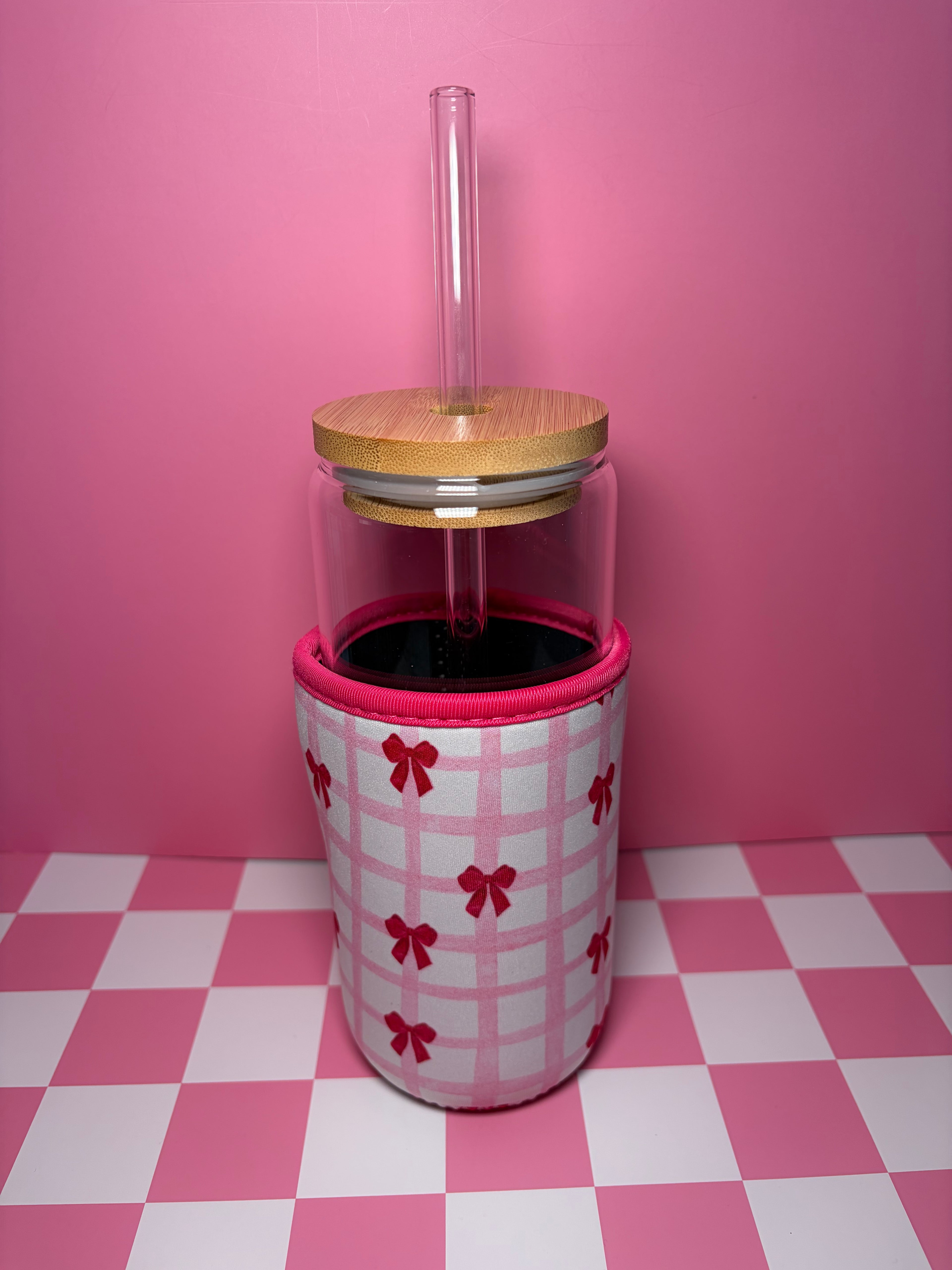Clear tumbler with wooden lid and pink bow-patterned sleeve on a pink checkered floor.
