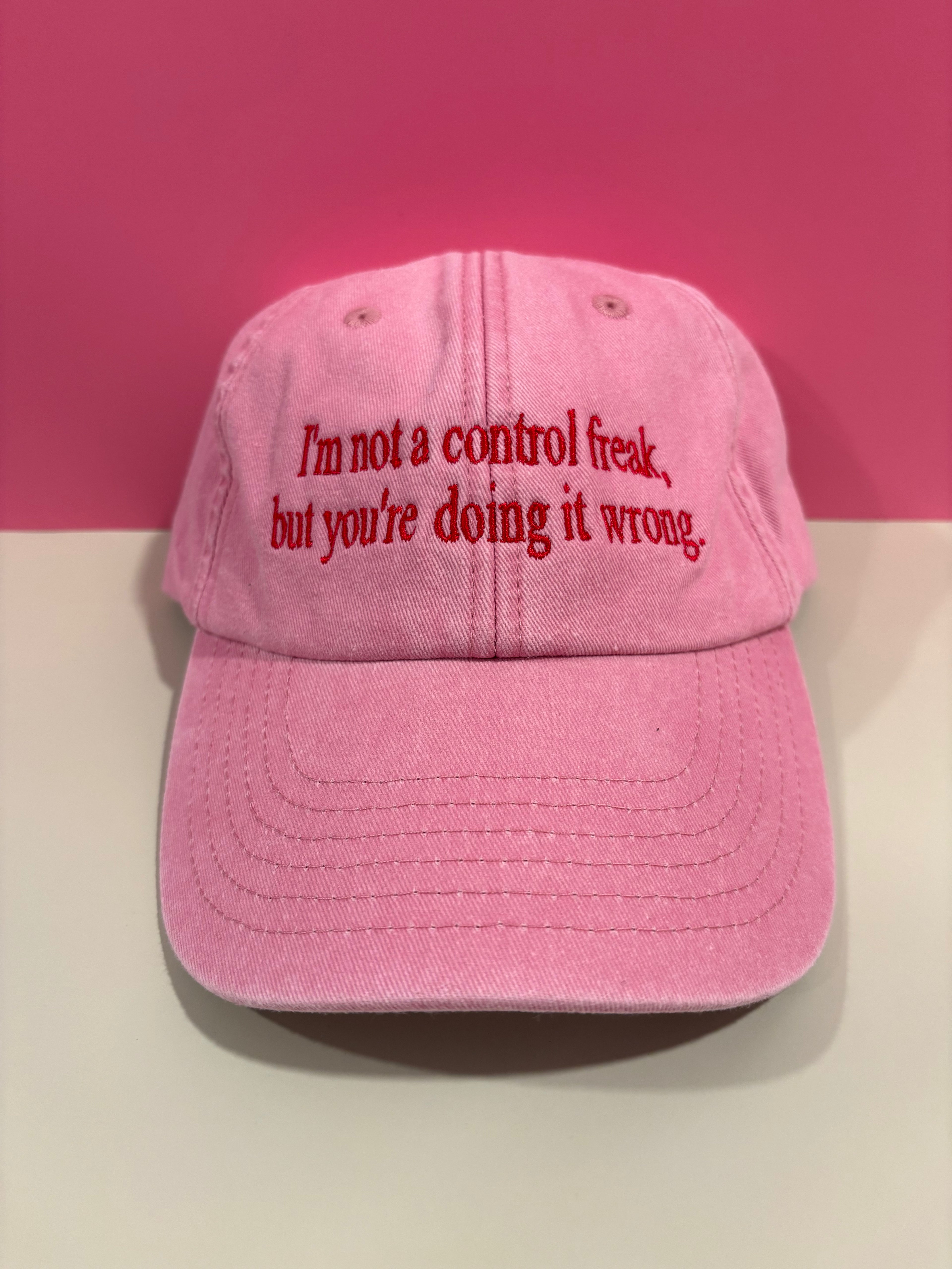 A pink baseball cap with red embroidered text reading 'I'm not a control freak, but you're doing it wrong.' displayed against a split pink and white background.