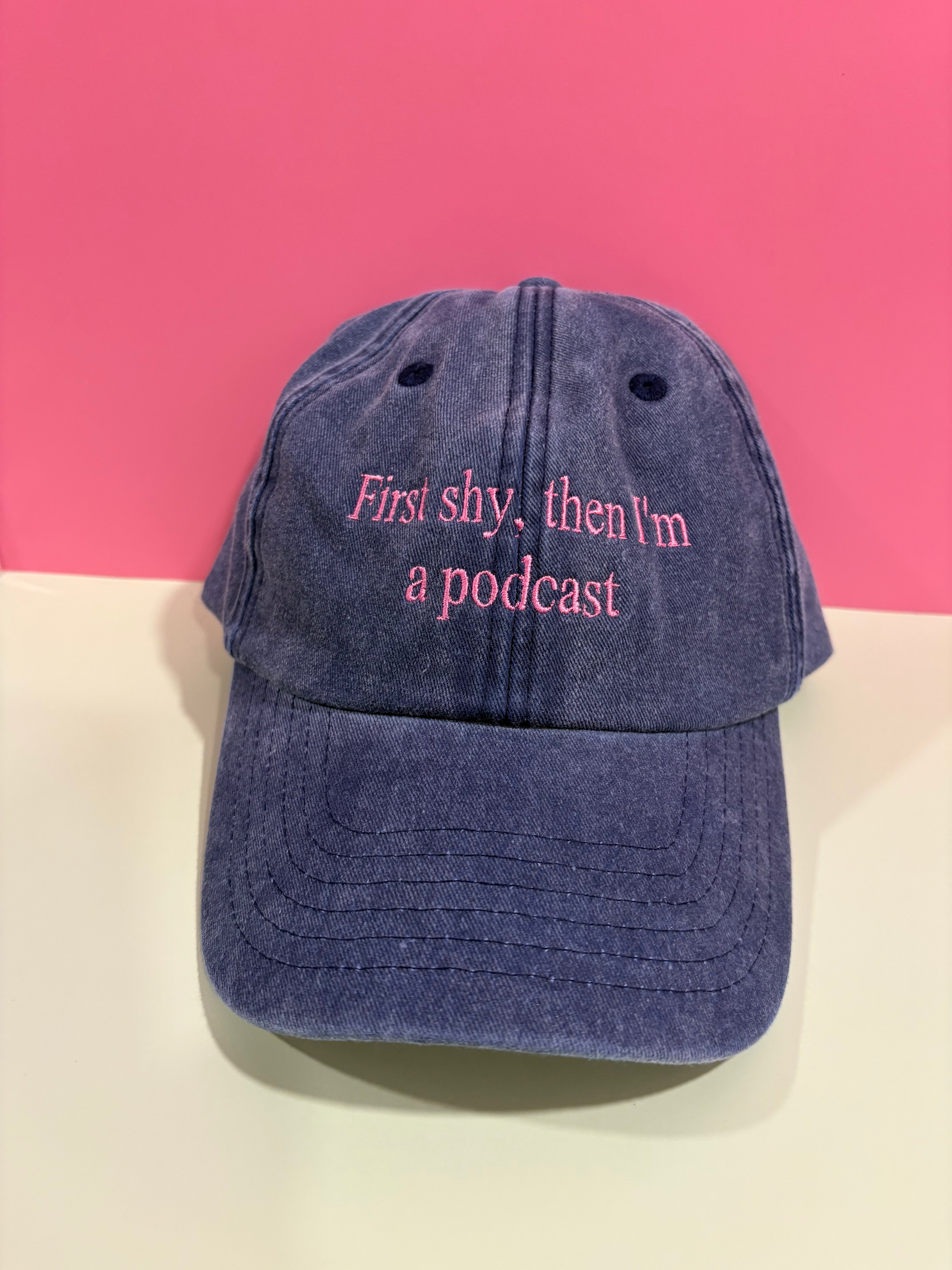 A gray denim baseball cap with pink embroidered text reading 'First shy, then I'm a podcast' on the front, displayed against a pink and cream background.