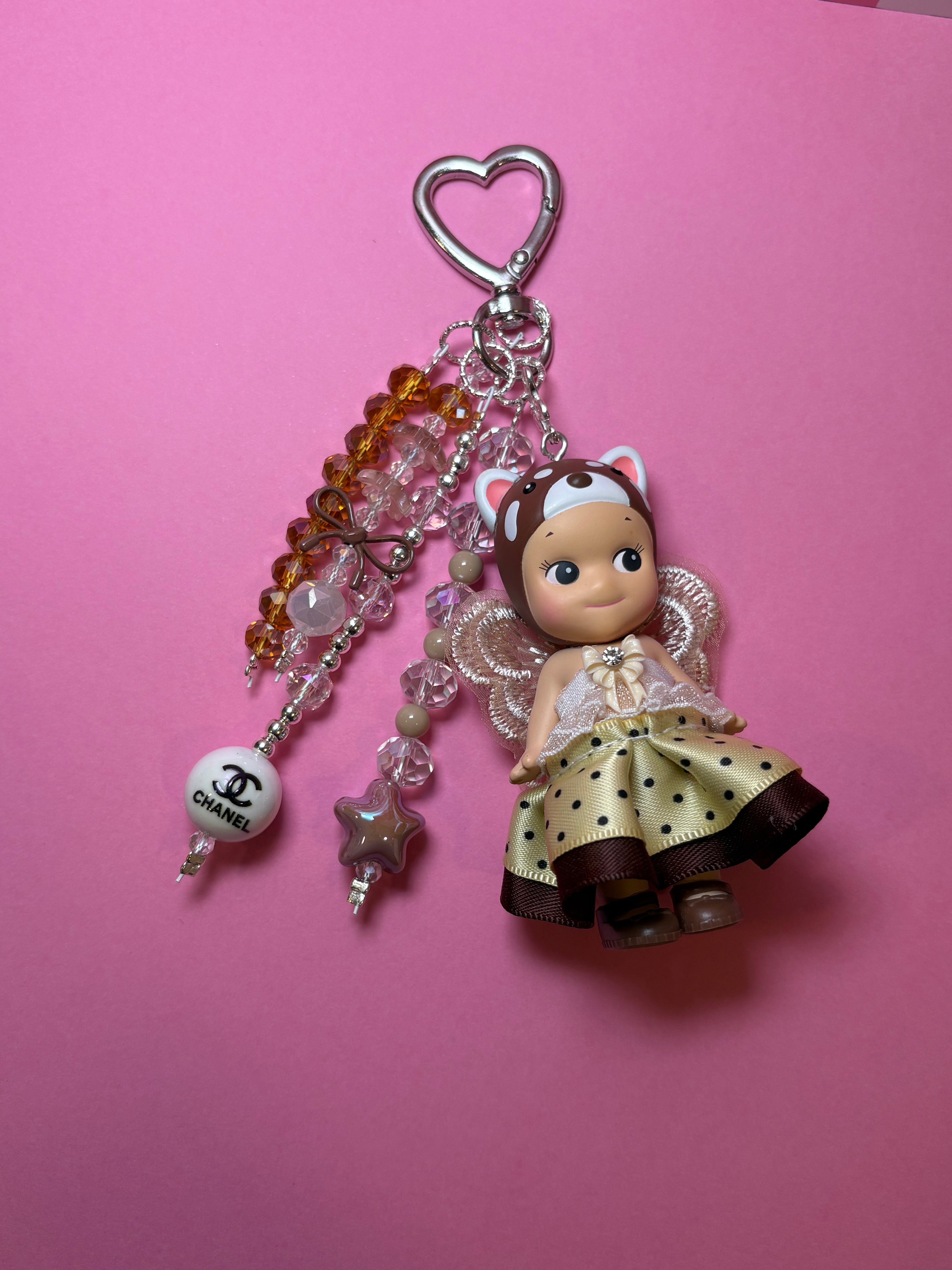 A keychain featuring a small lesser panda sonny angel doll with a cream and brown polka-dot dress, and cat-ear headband, hanging from a silver heart-shaped clasp with clear beads, amber beads, and a white Chanel logo bead on a solid pink background.