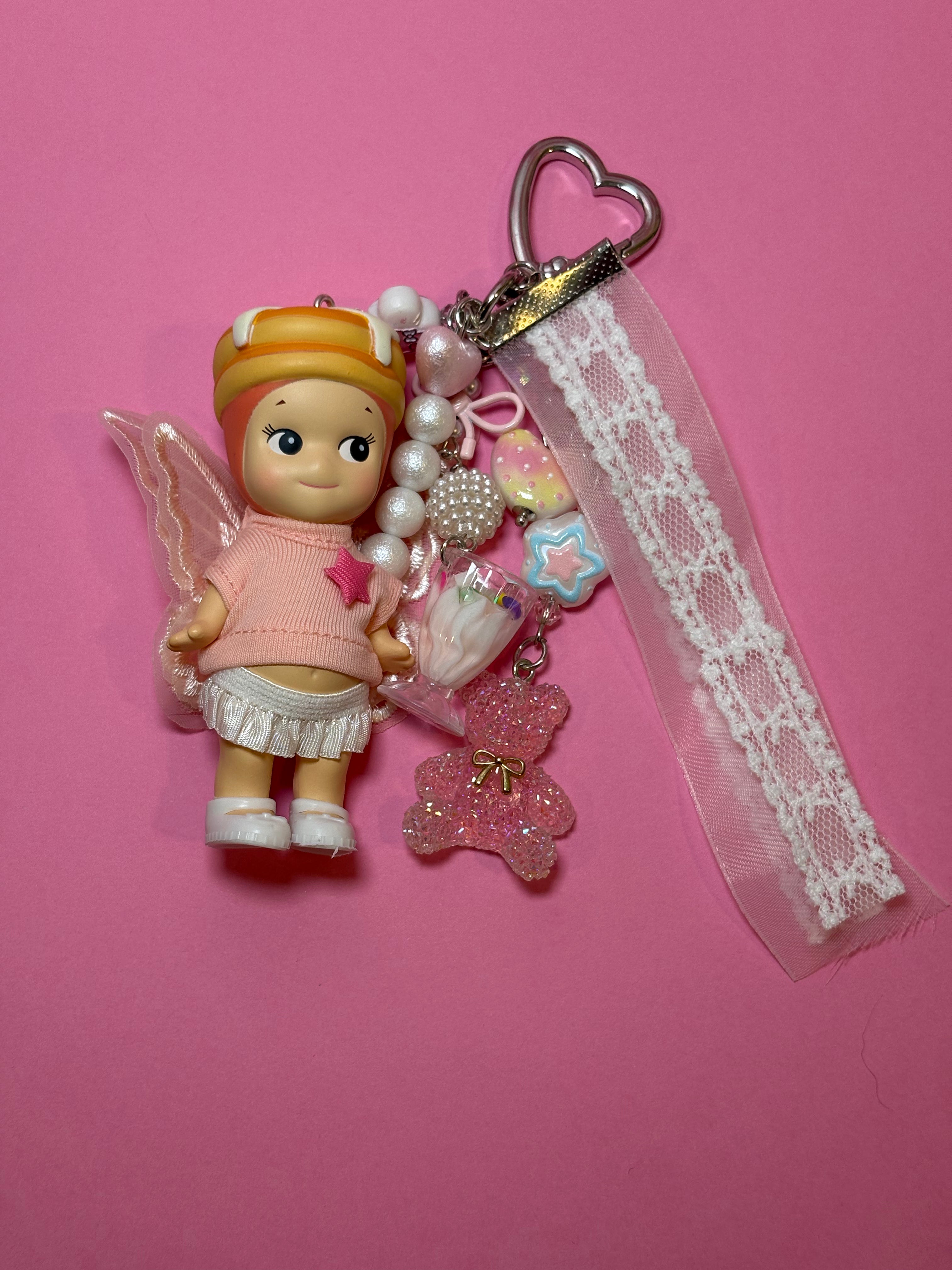 A cupcake sonny angel charm keychain with white lace ribbon, pearl beads, and a silver heart clasp, set against a solid pink background.