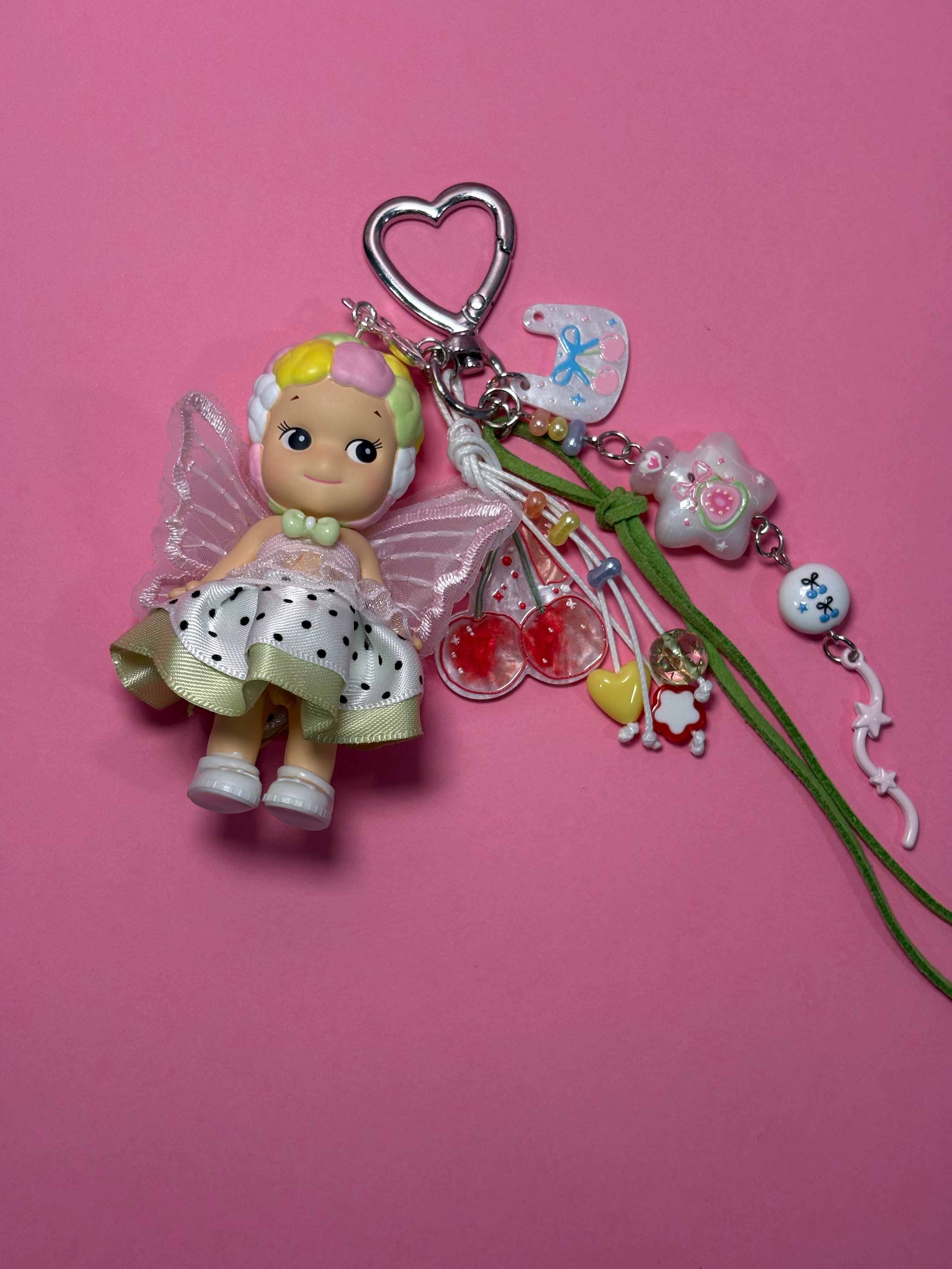 A colorful konpeito sonny angel keychain with translucent wings, a silver heart-shaped clasp, and multiple dangling charms including a blue bird, cherry, star, and green cord details, set against a solid pink background.