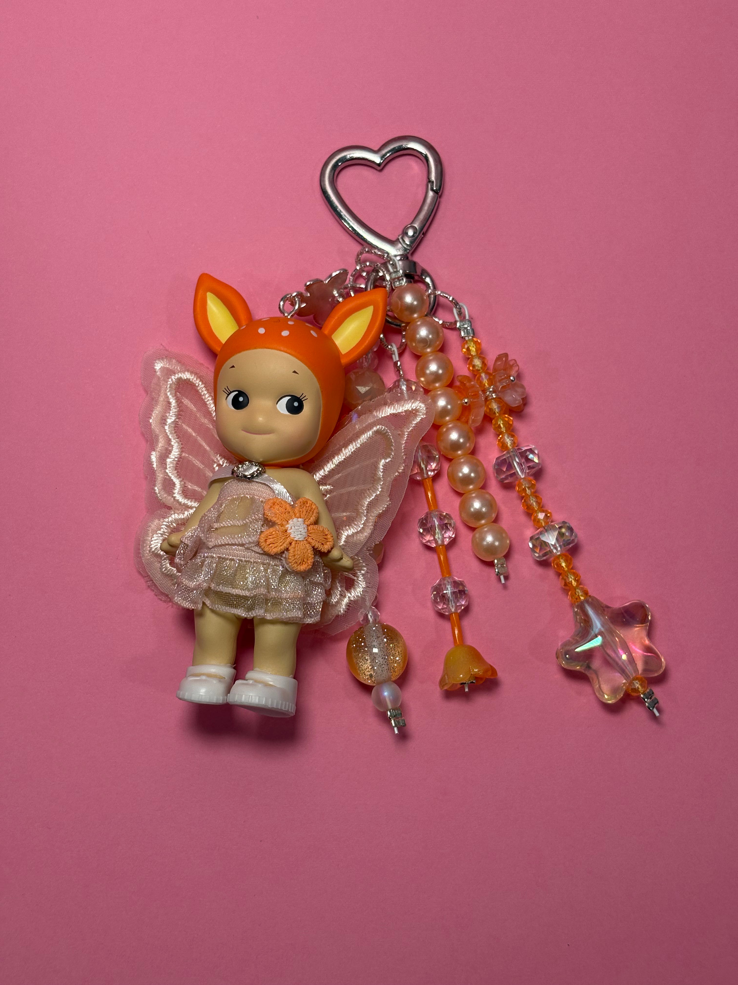A cute keychain charm featuring a fawn sonny angel with translucent wings, and a beige dress, hanging from a silver heart-shaped clasp with dangling beaded strands on a solid pink background.