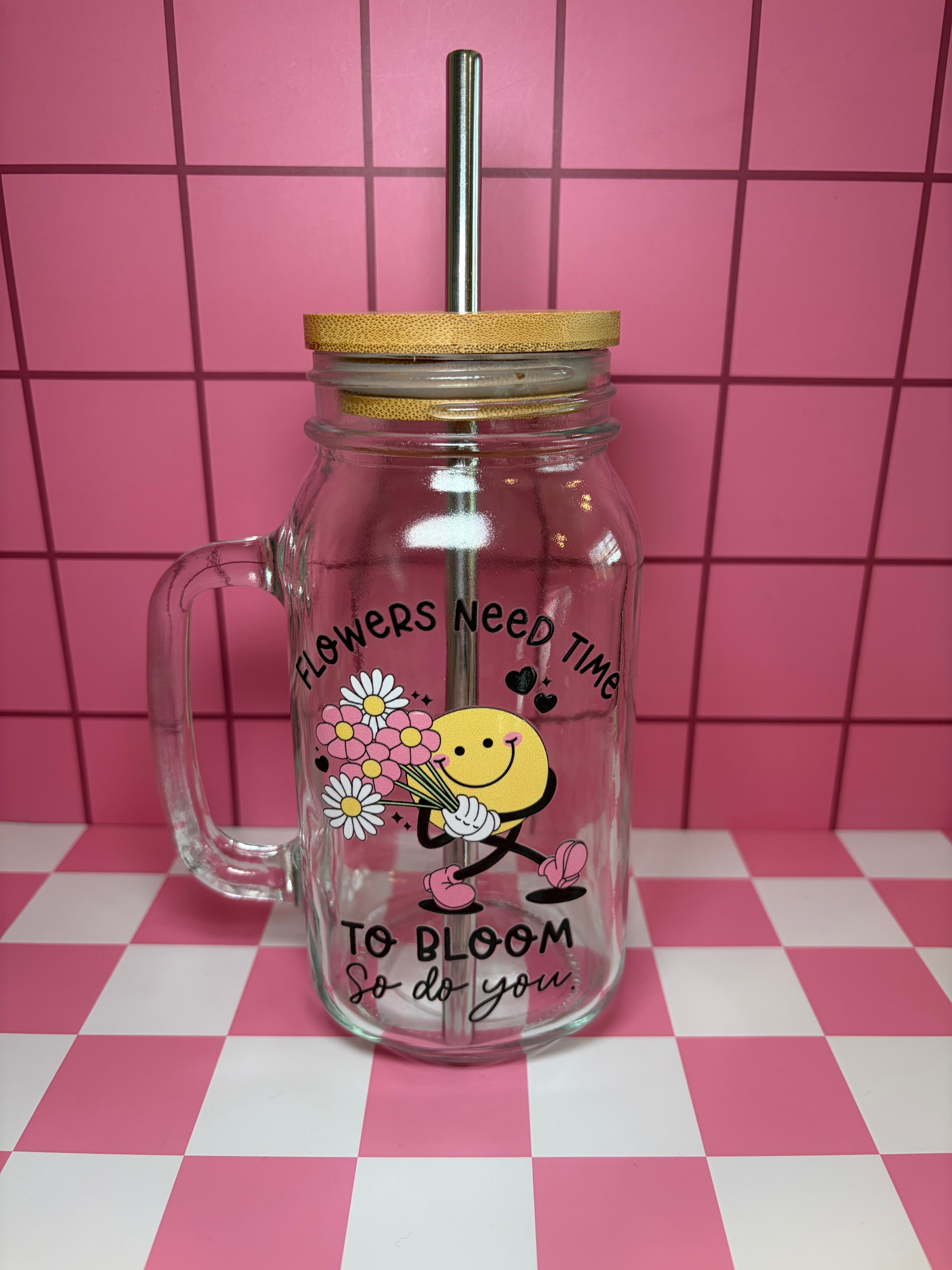 Clear glass jar with handle and smiley face holding flowers with "flowers need time to bloom, so do you" text
