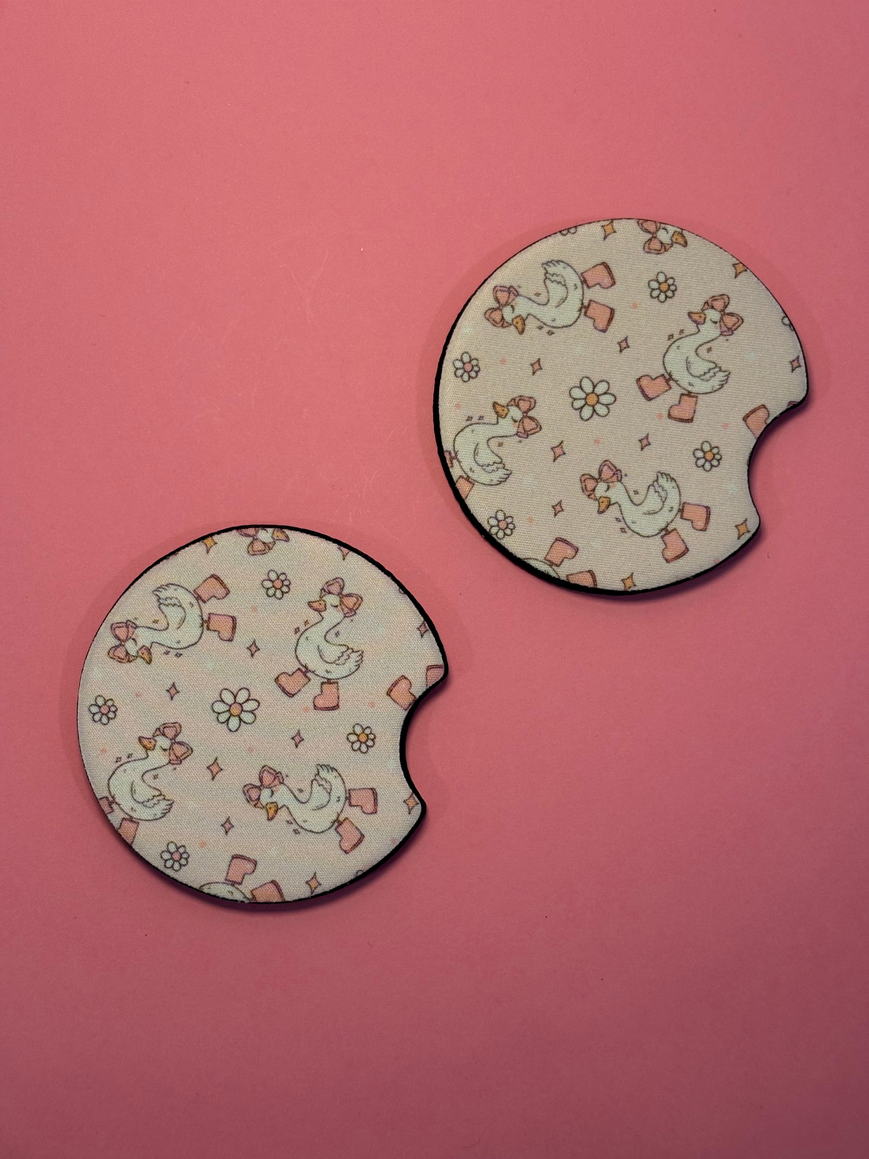 Car coasters with a beige background, printed with a cute pattern of white silly geese wearing pink bows and small flowers, set against a solid pink background.