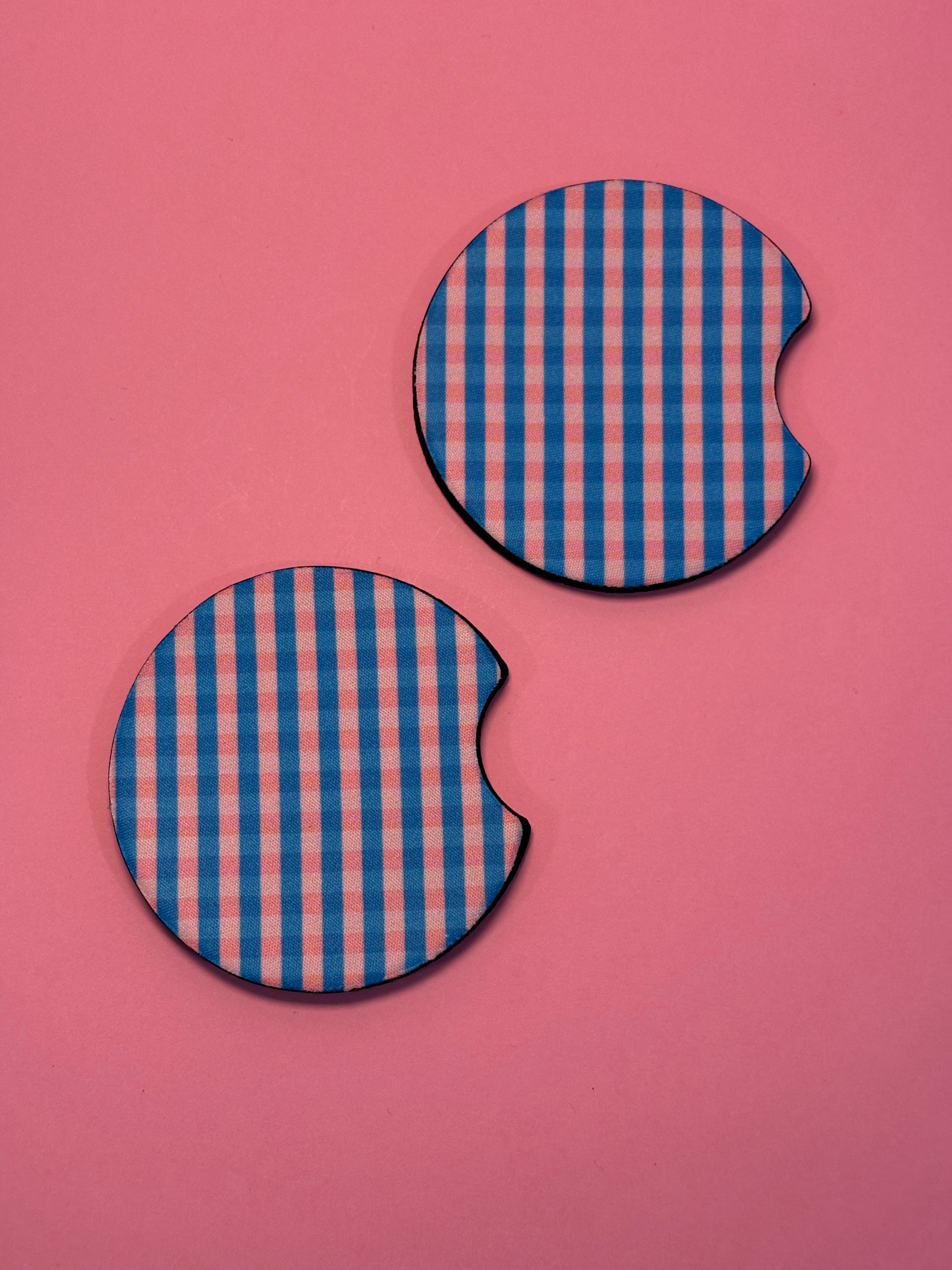 Two circular car coasters with a blue and pink gingham checkered pattern, featuring a small cutout on one side, displayed on a solid pink background.