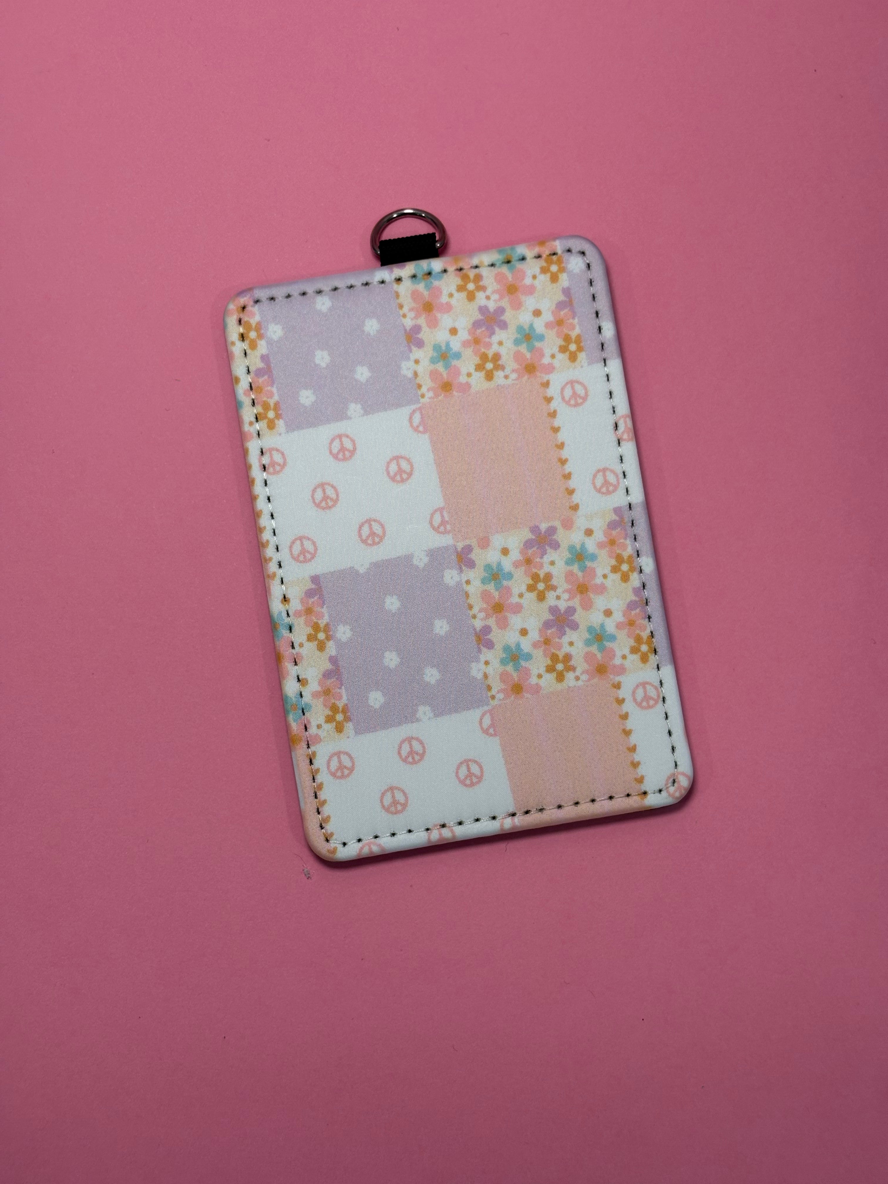 A rectangular patchwork-style card holder/ID badge case with a small metal ring at the top, shown on a solid pink background. The case features a retro floral and peace-sign patchwork pattern in pastel lavender, peach, white, and orange.