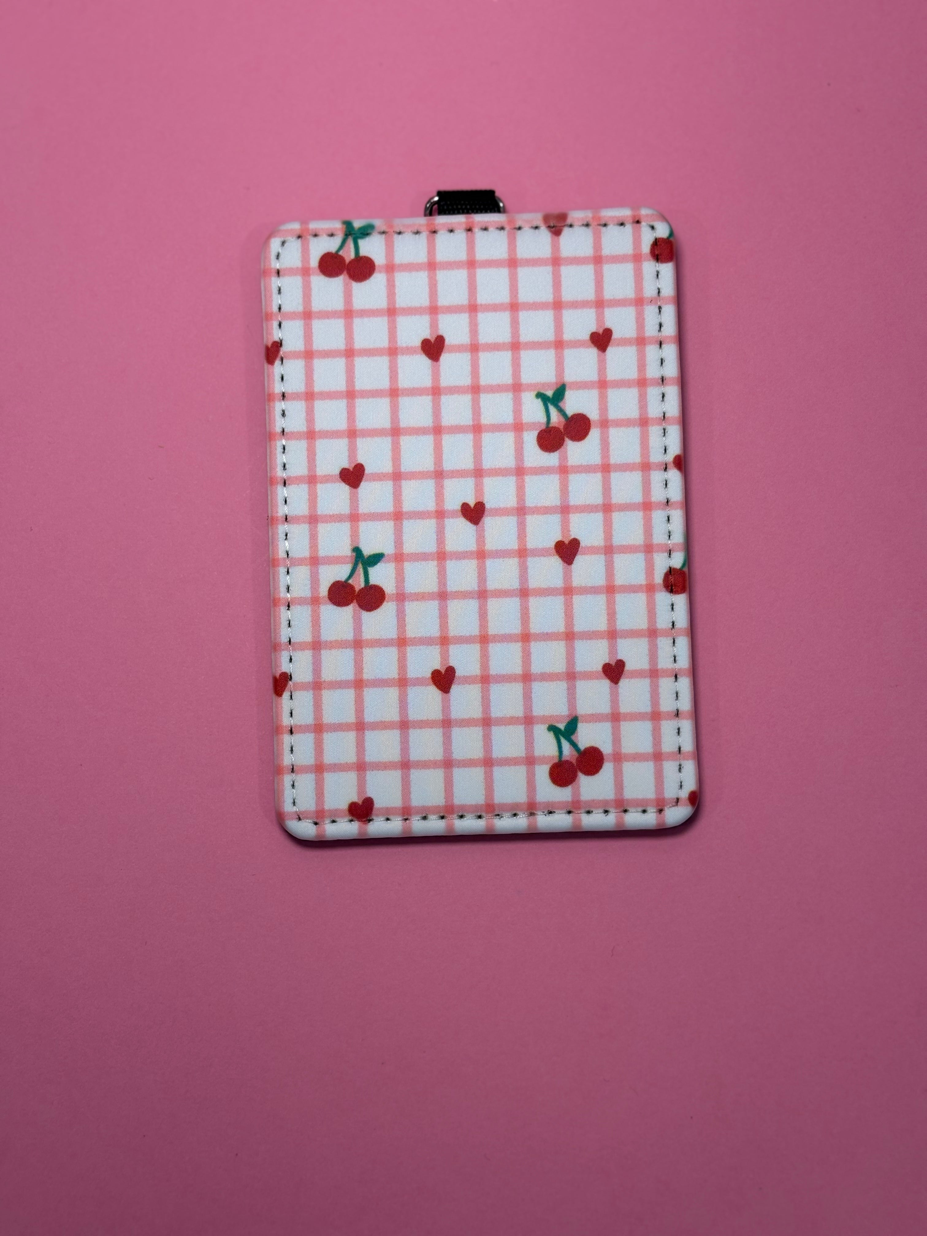 A rectangular ID badge holder with a pink and white gingham pattern, decorated with red cherries and small red hearts, shown on a solid pink background.