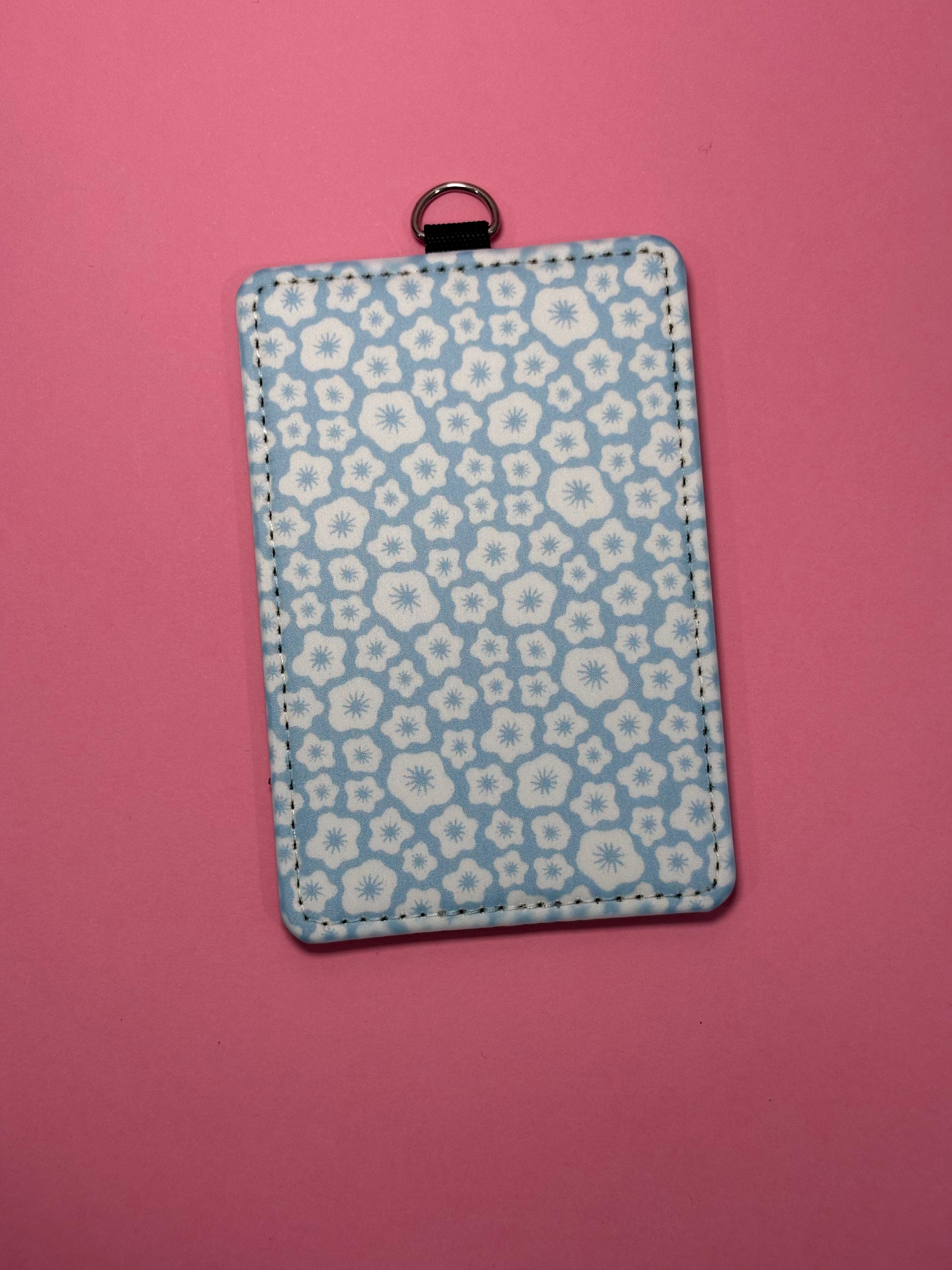 A rectangular ID badge/card holder with a light blue and white floral pattern, featuring a small black metal ring at the top, set against a solid pink background.