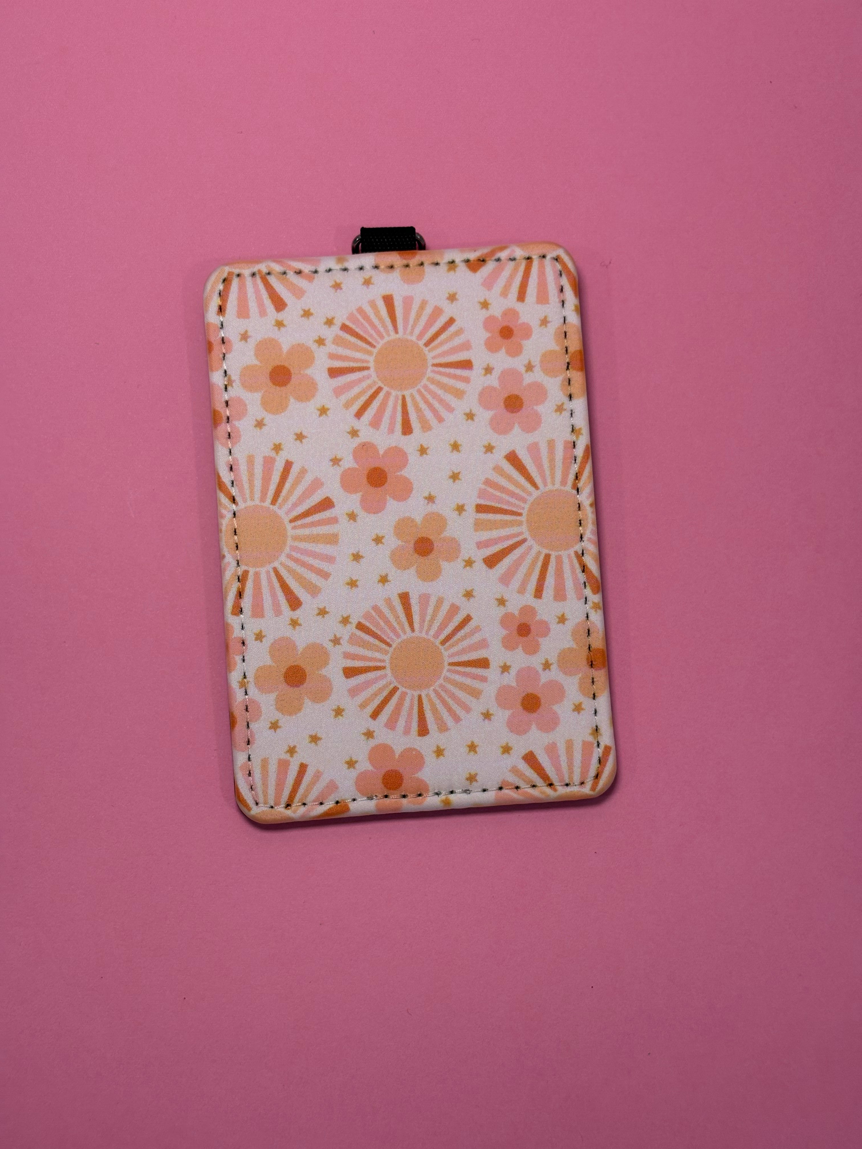 A rectangular ID badge holder with a retro floral pattern in orange, peach, and yellow on a white background, featuring a black plastic top loop, set against a solid pink background.