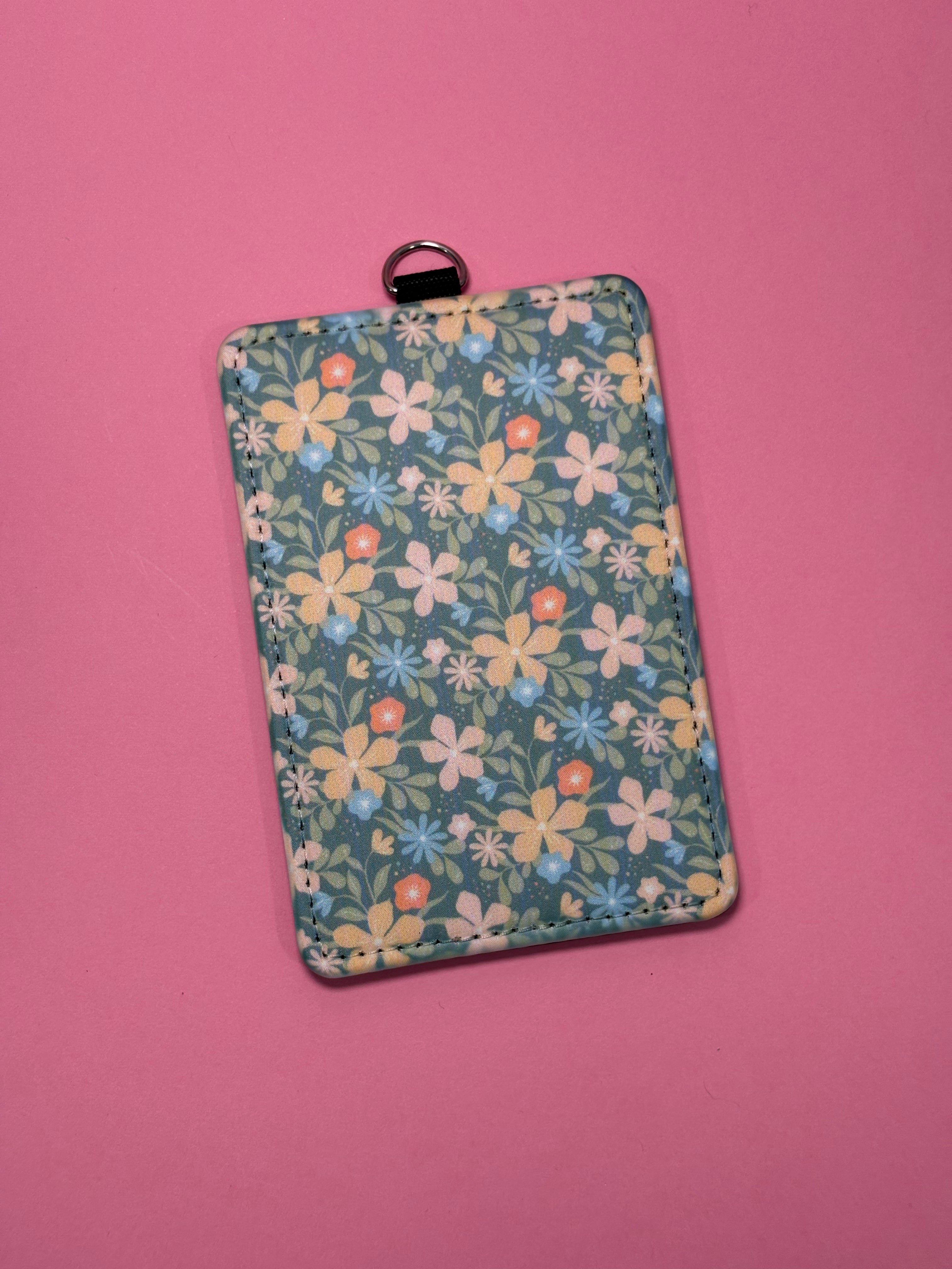 A rectangular card holder/ID badge sleeve with a sage floral pattern, featuring a small silver metal ring at the top, set against a solid pink background.