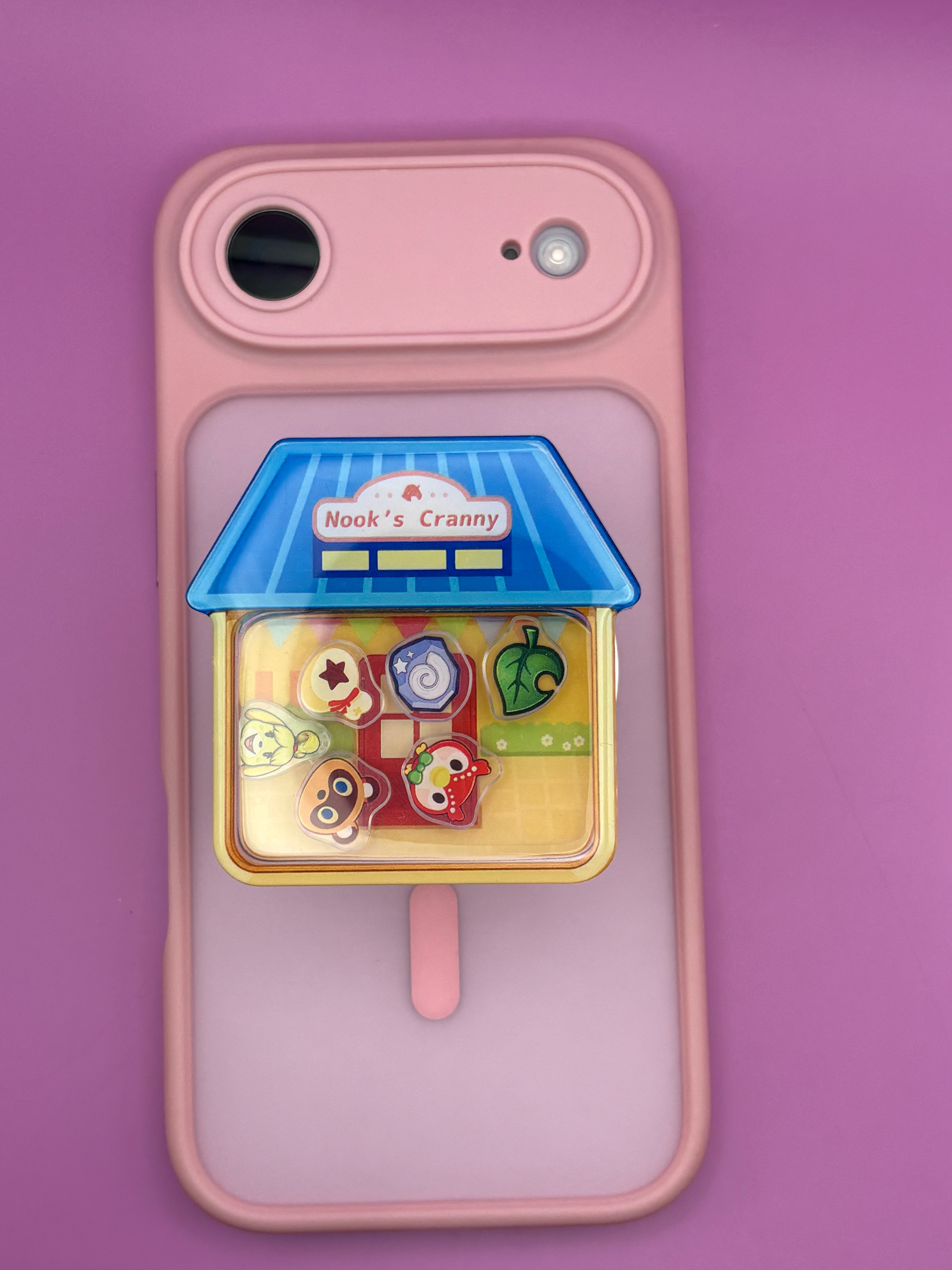 A pink phone case with a built-in transparent panel showing a small toy shop charm labeled 'Nook's Cranny' on a purple background.