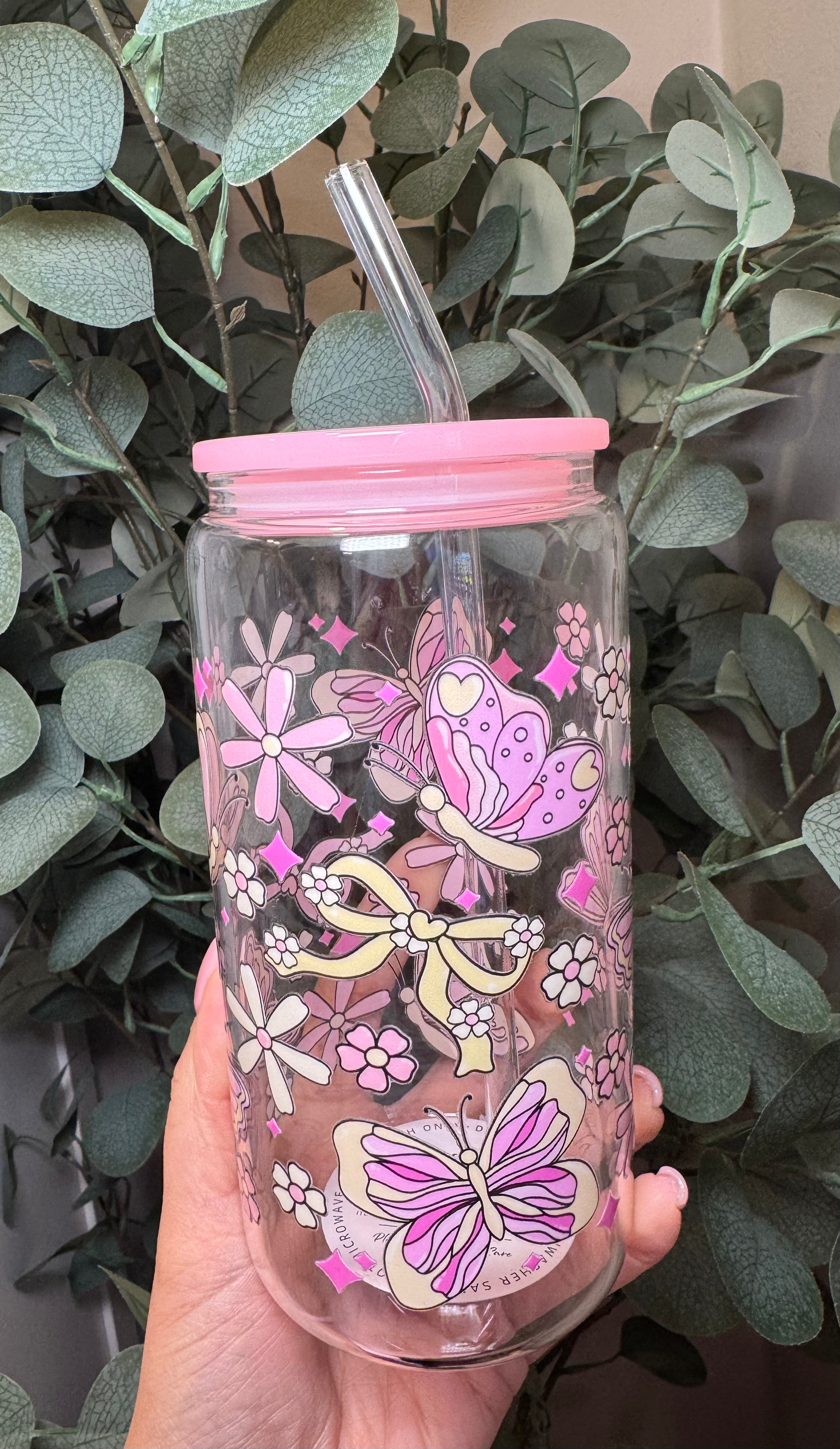 Clear tumbler with pink lid and pink and beige butterfly and bow design held in front of green leaves