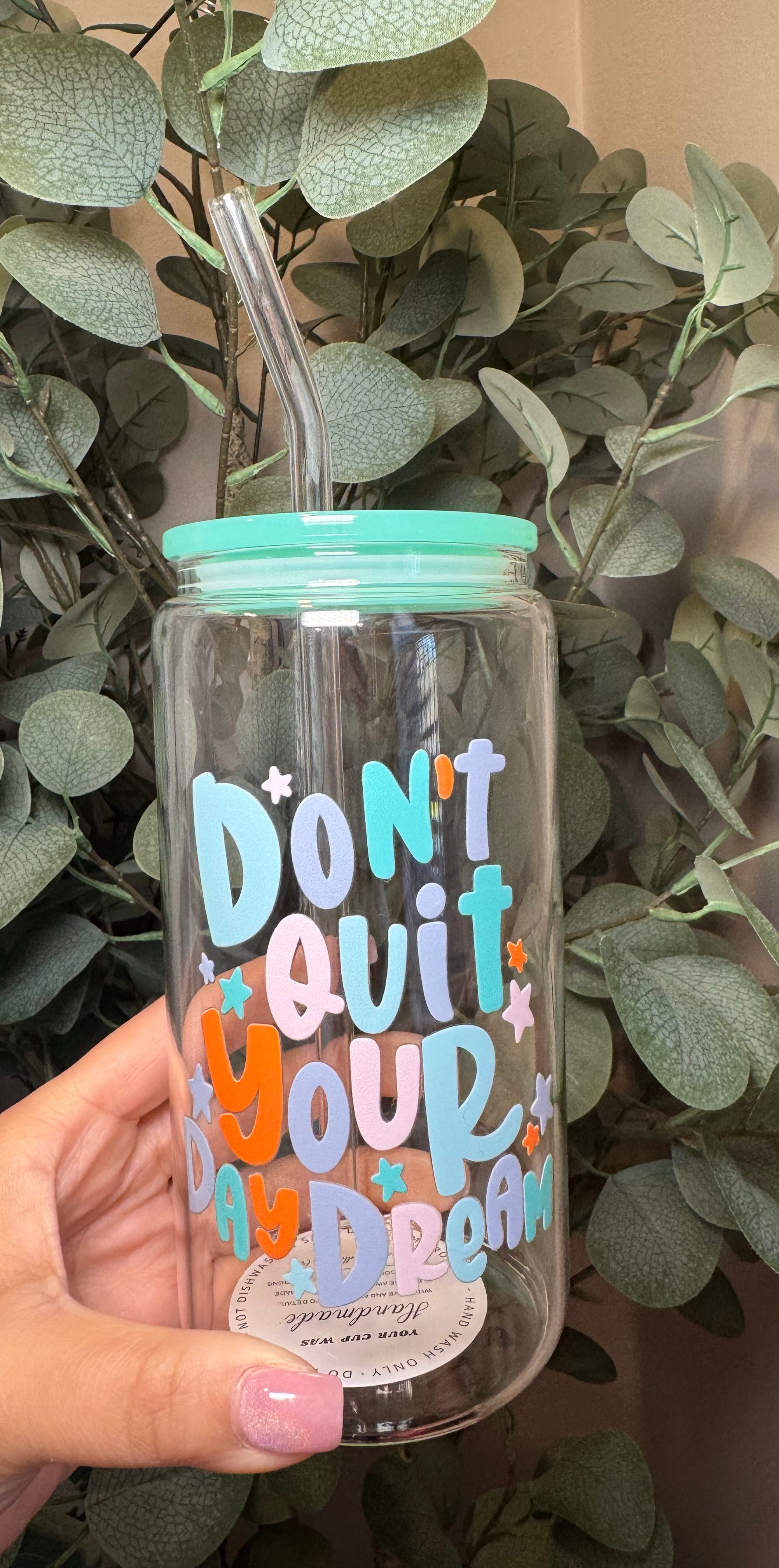 Clear tumbler with a teal lid and colorful text "Don't Quit Your Day Dream" and a straw, held in front of green foliage.