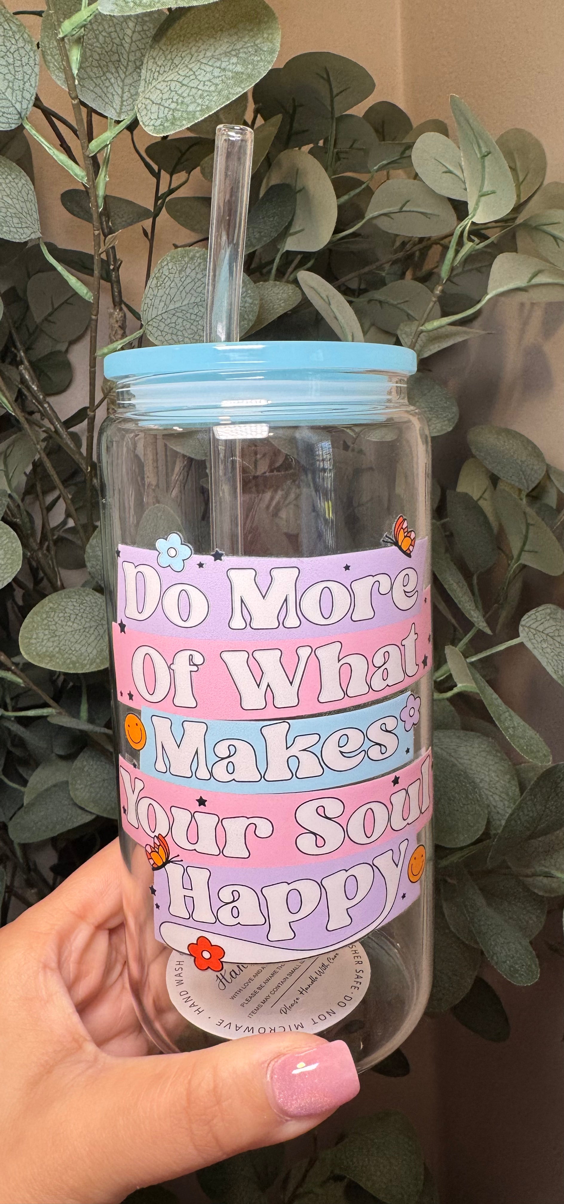 Clear tumbler with colorful text "Do More of What Makes Your Soul Happy" and a straw, held in front of green leaves.