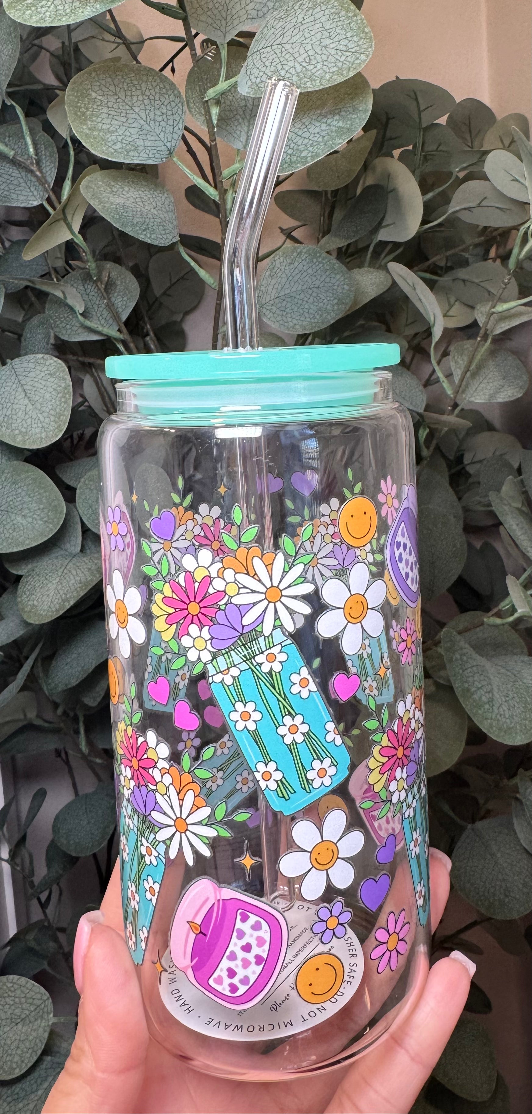 Clear tumbler with floral design and turquoise lid held in front of green foliage