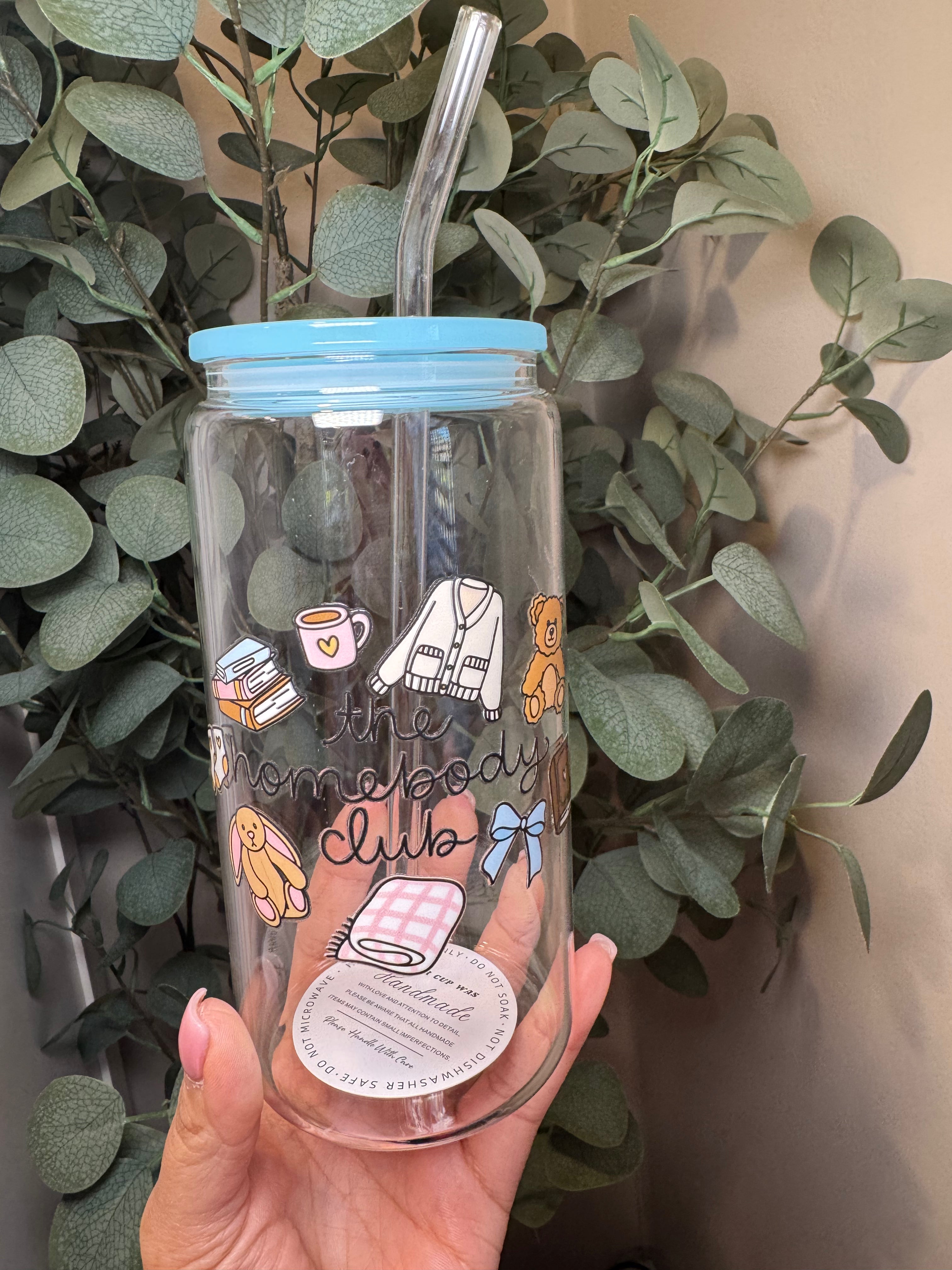 Clear tumbler with colorful designs and text "The Homebody Club" held in front of green leaves