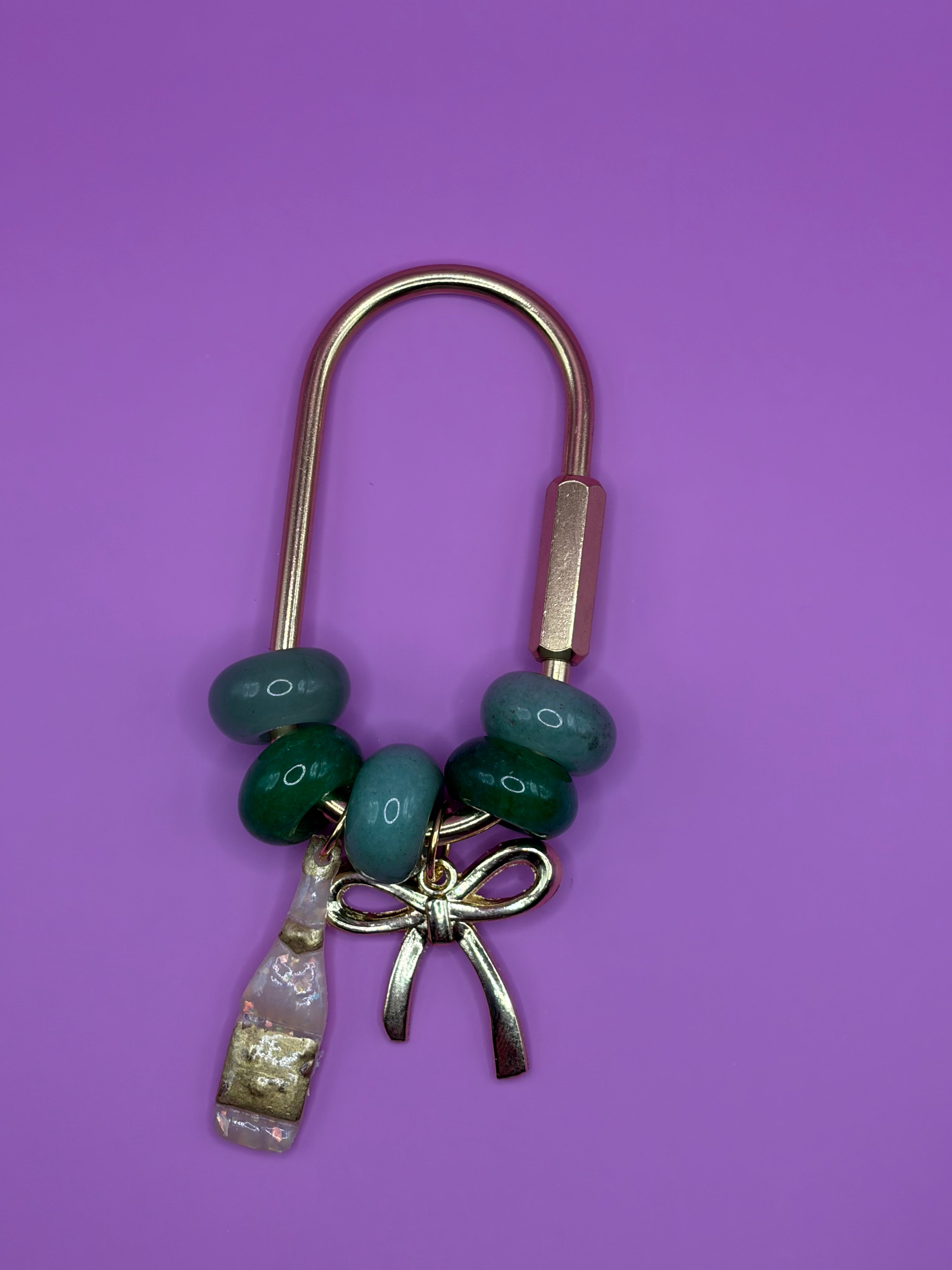 A gold-tone metal keychain or bookmark featuring dark green and teal beads, a small metallic bow charm, and a clear glass bottle charm filled with gold glitter, set against a solid purple background.