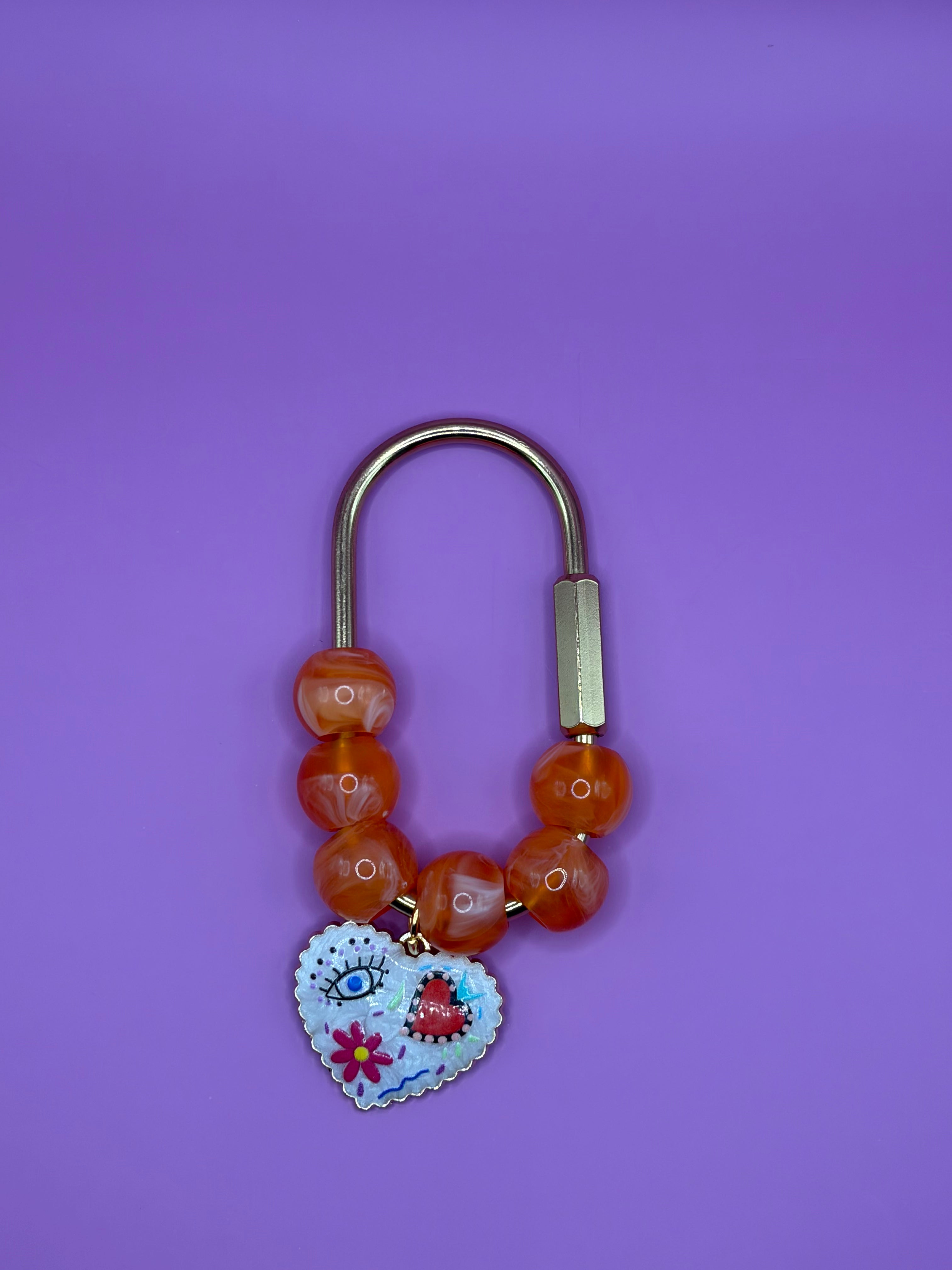 A beaded bracelet with orange agate-like beads, a silver-toned metal clasp, and a decorative heart charm featuring a colorful face and floral details, set against a solid purple background.