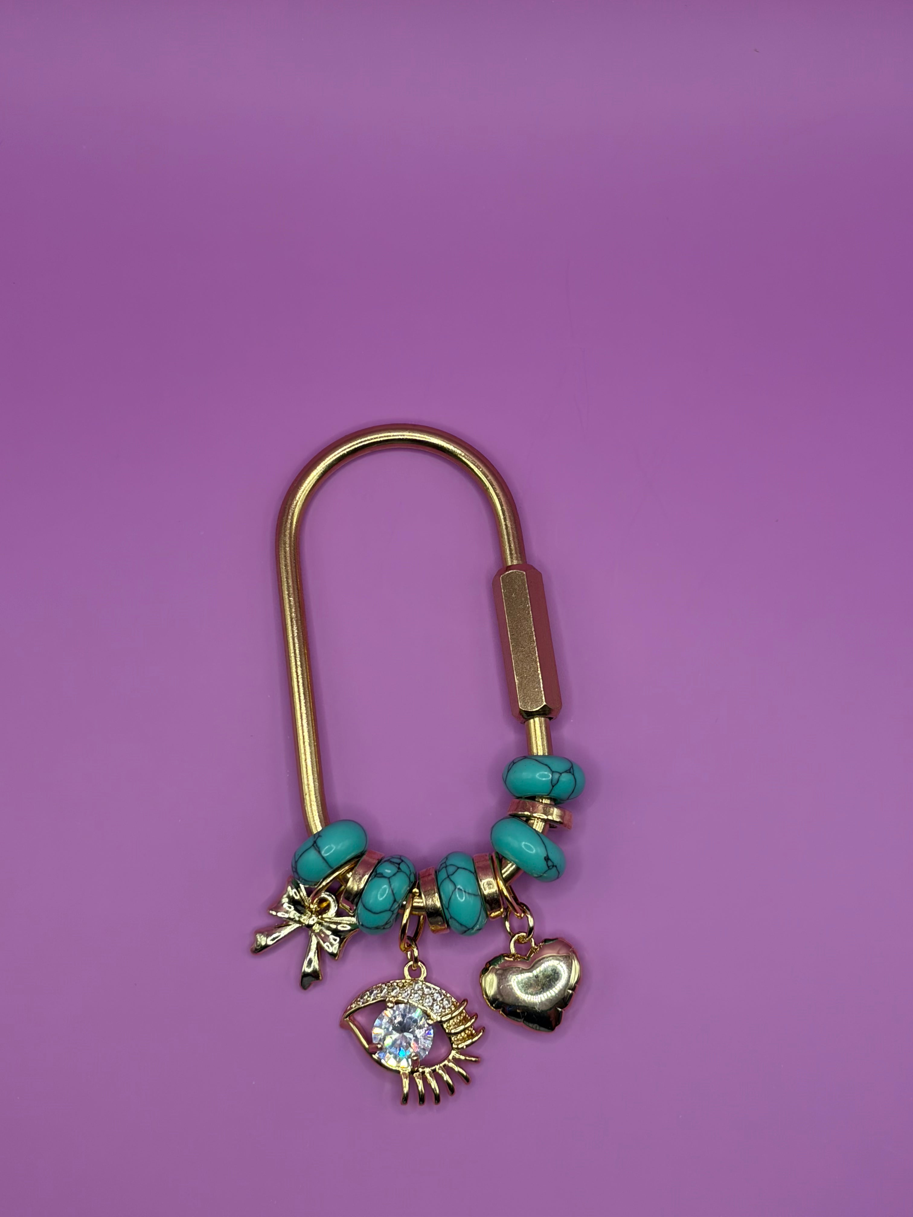 A gold-tone charm keychain featuring turquoise beads, a small angel charm, a heart-shaped charm, and an eye of providence charm, displayed on a solid purple background.