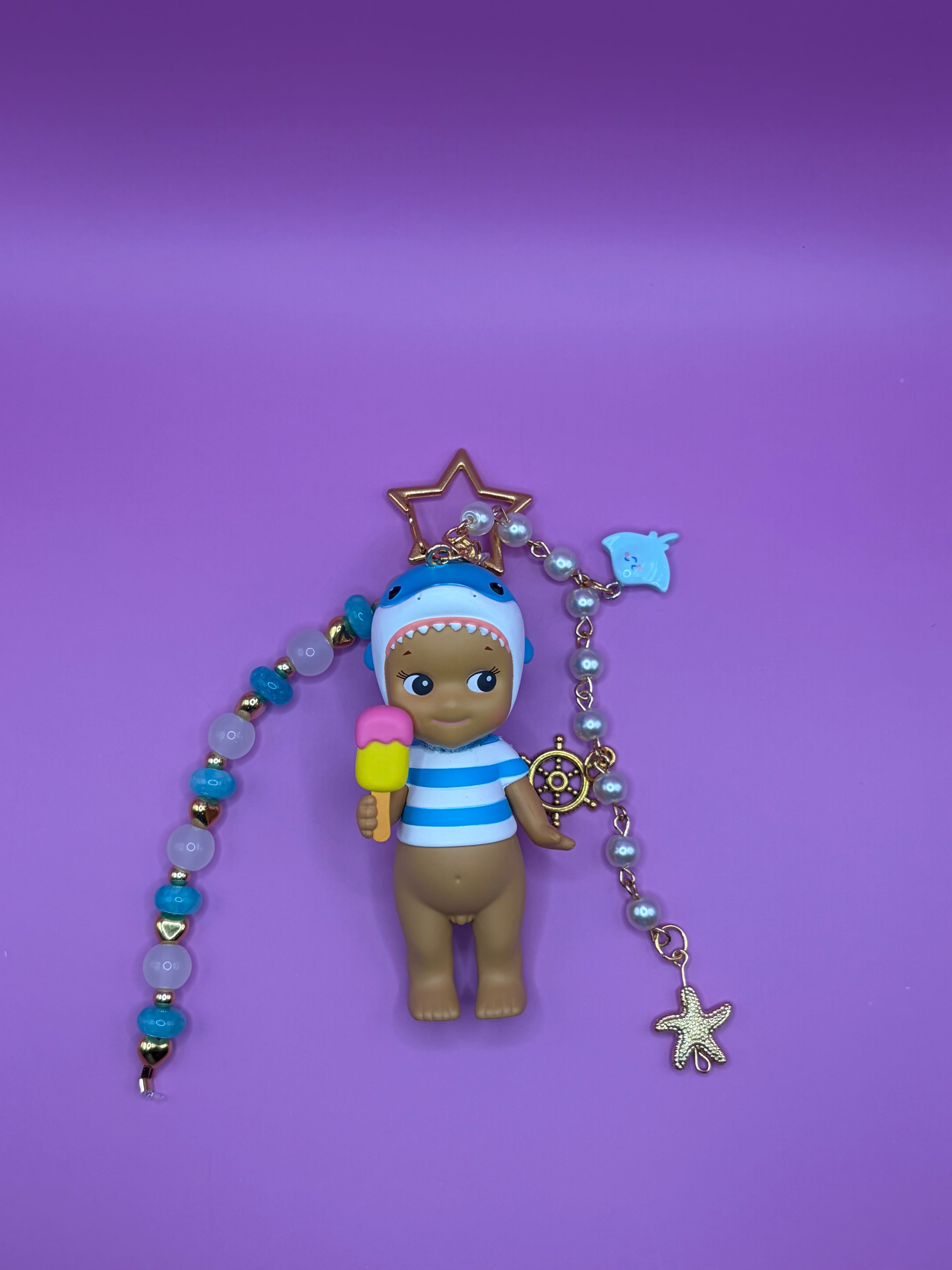 A small doll figurine with brown skin, wearing a blue and white striped shirt and a blue hat, holding a pink and yellow ice cream cone. The doll is attached to a beaded charm bracelet with gold-toned accents, including a starfish, ship's wheel, and blue fish charms, set against a solid purple background.