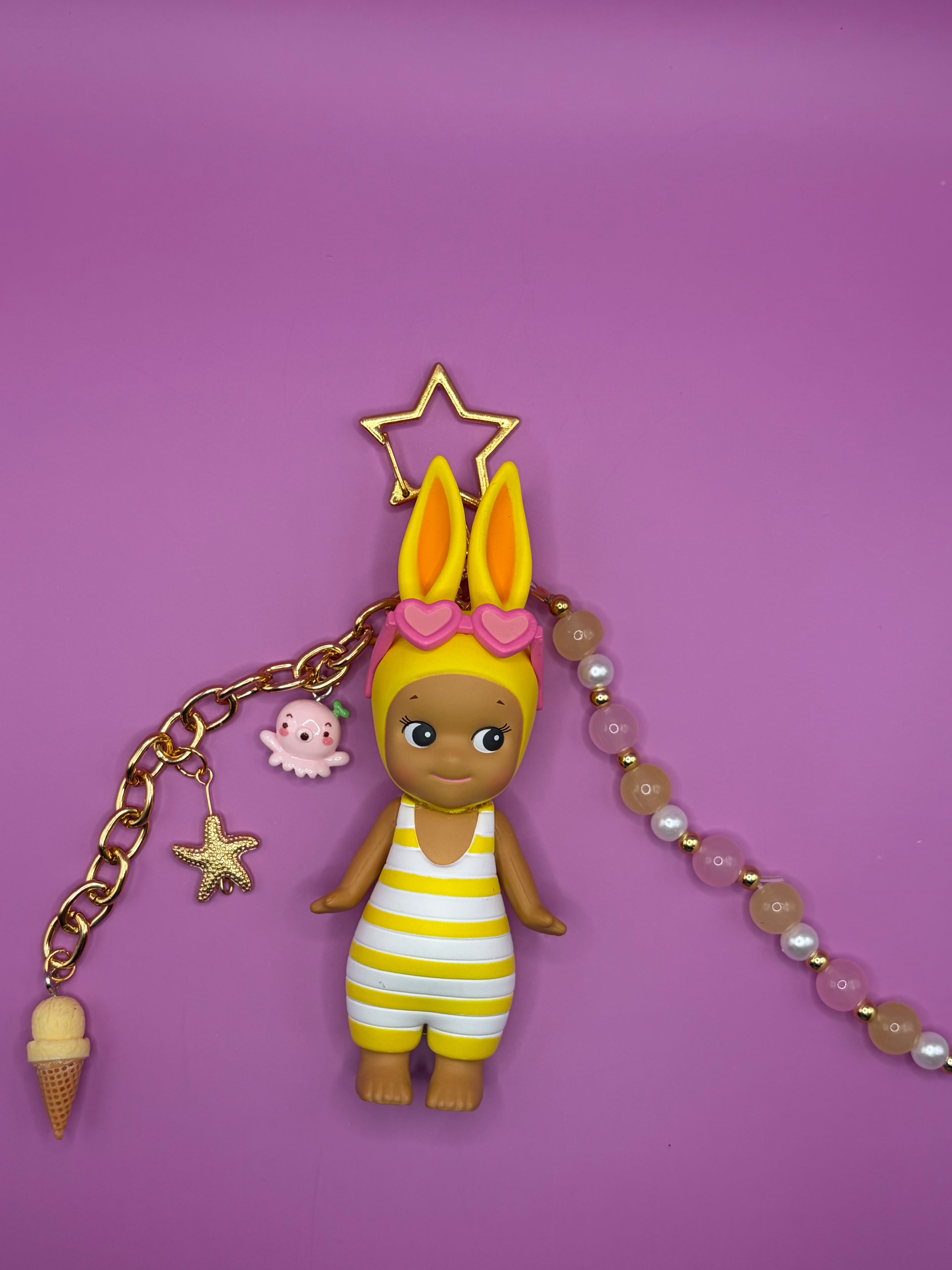 A doll charm with yellow bunny ears, a pink heart headband, and a yellow-and-white striped romper, attached to a gold-tone chain with star and octopus charms, a beige ice cream cone, and a pink-and-white beaded strap, displayed on a solid purple background.