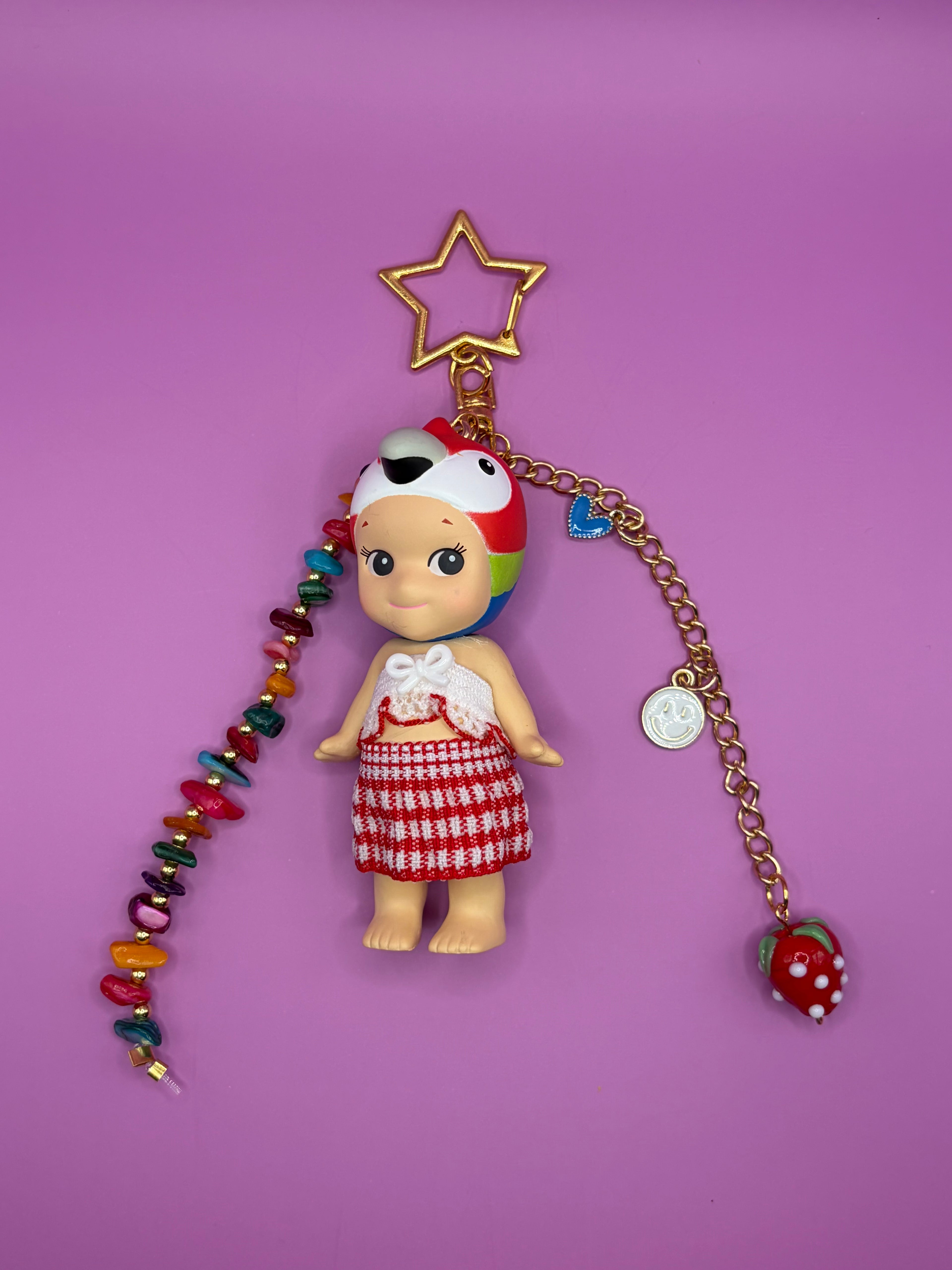 A doll keychain featuring a small doll with a red and white checkered skirt, a colorful striped hat, and a gold star-shaped keyring, set against a solid purple background.