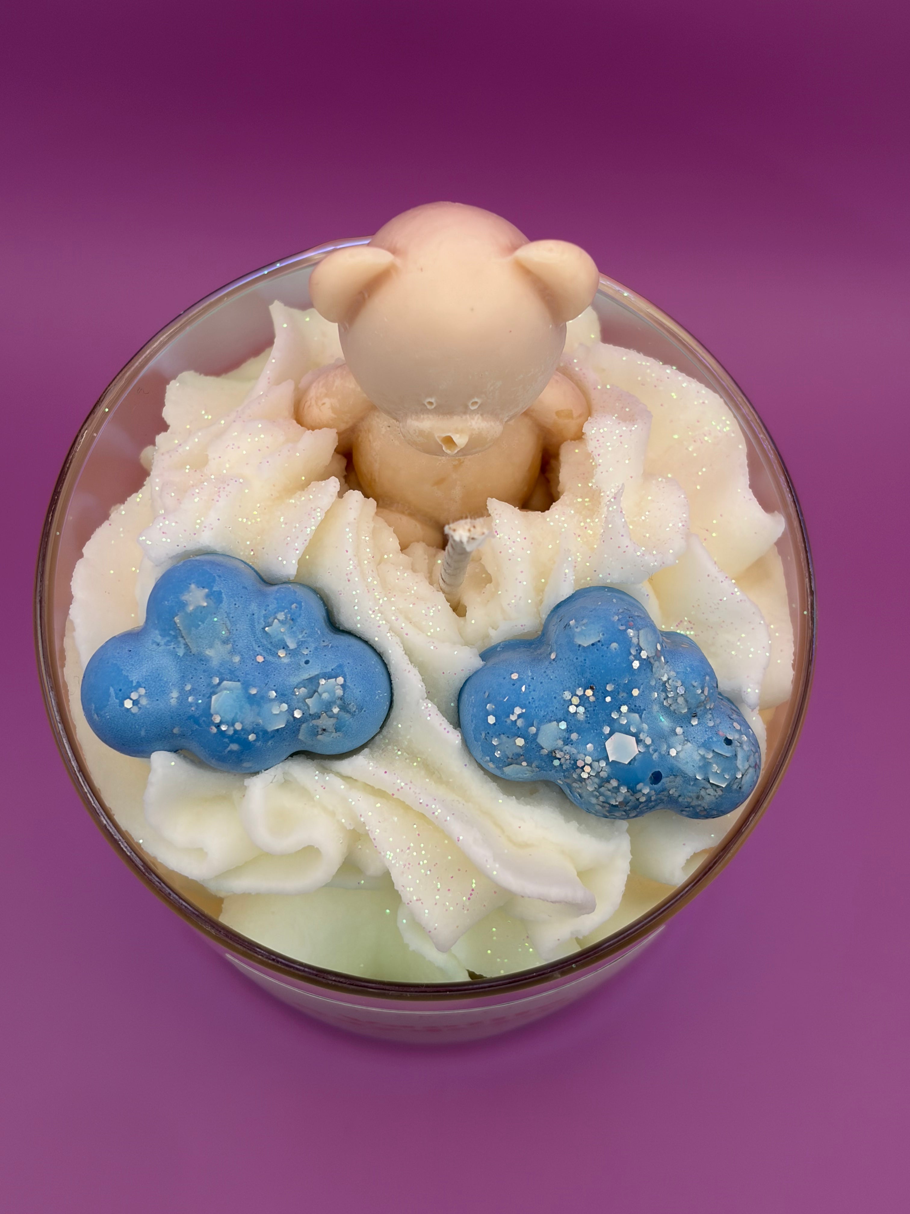A candle topped with white whipped cream, two blue cloud-shapes with white sprinkles, and a small beige bear figurine on top, set against a solid purple background.