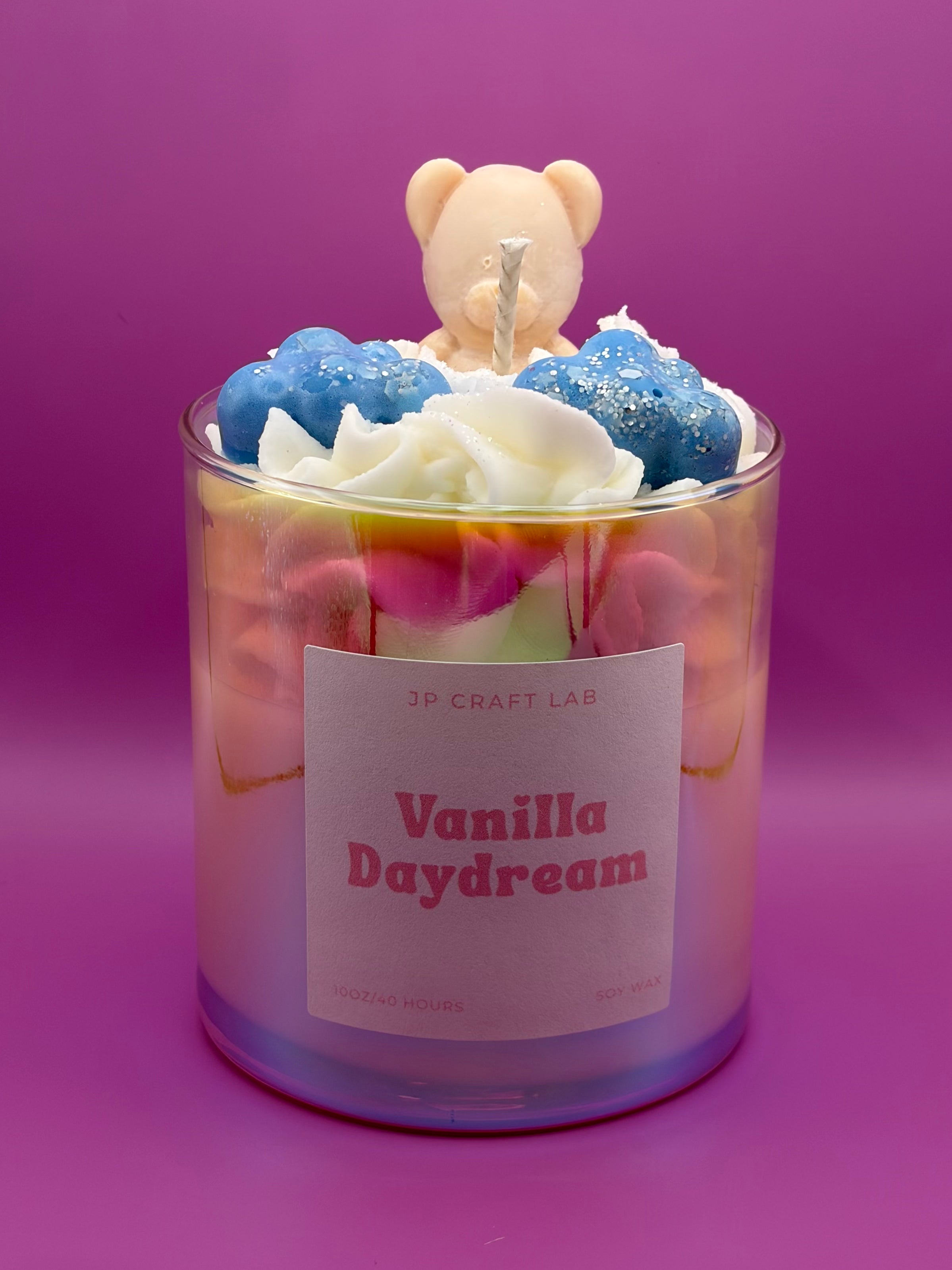 A jar candle labeled 'Vanilla Daydream' by JP Craft Lab, filled with layered pastel wax and topped with blue clouds, whipped wax, and a small teddy bear figure, set against a solid purple background.