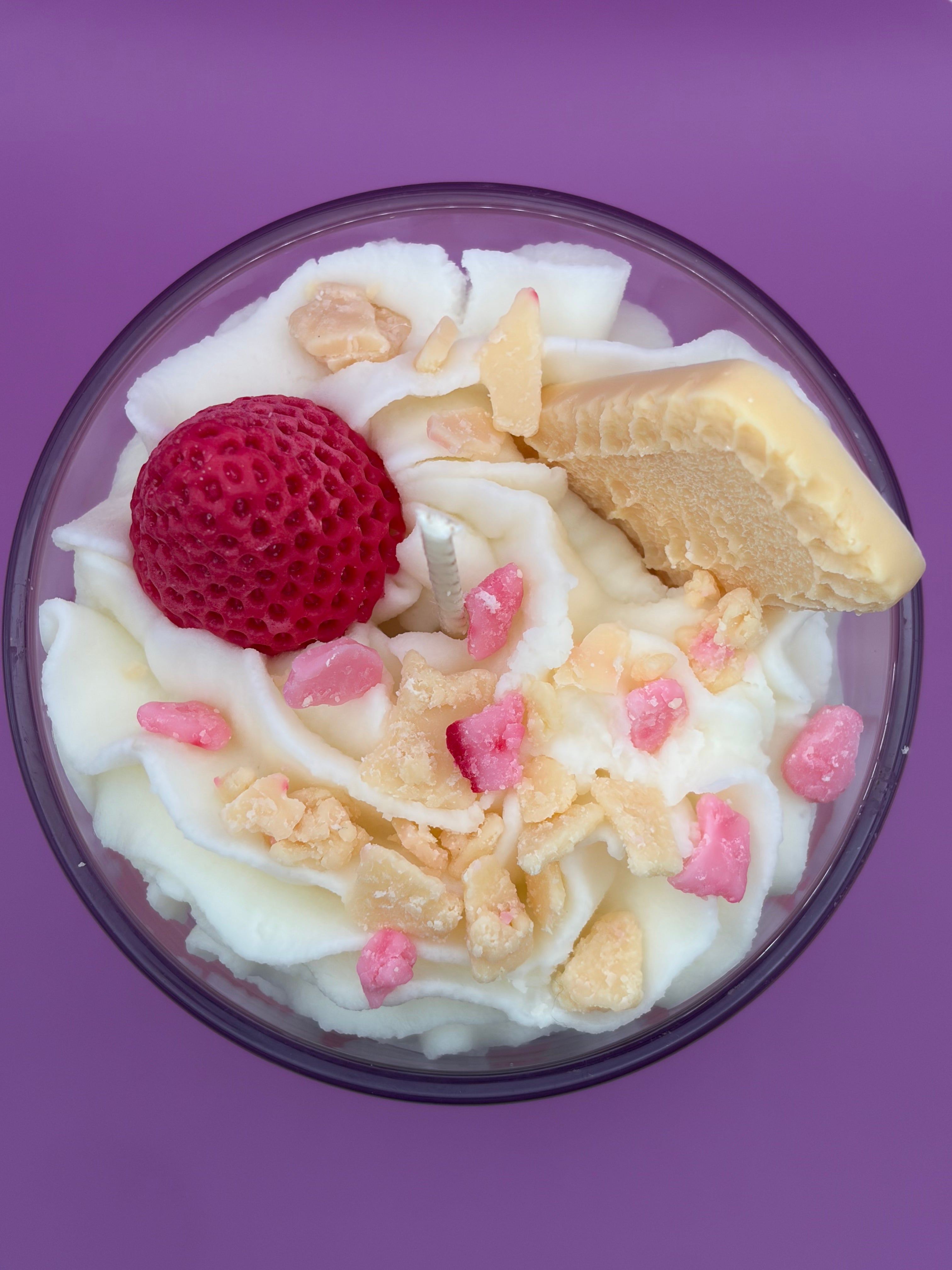 A top-down view of a dessert candle in a clear glass bowl, featuring whipped cream, a red strawberry, a piece of cream cheese, and pink and beige candy pieces on a solid purple background.