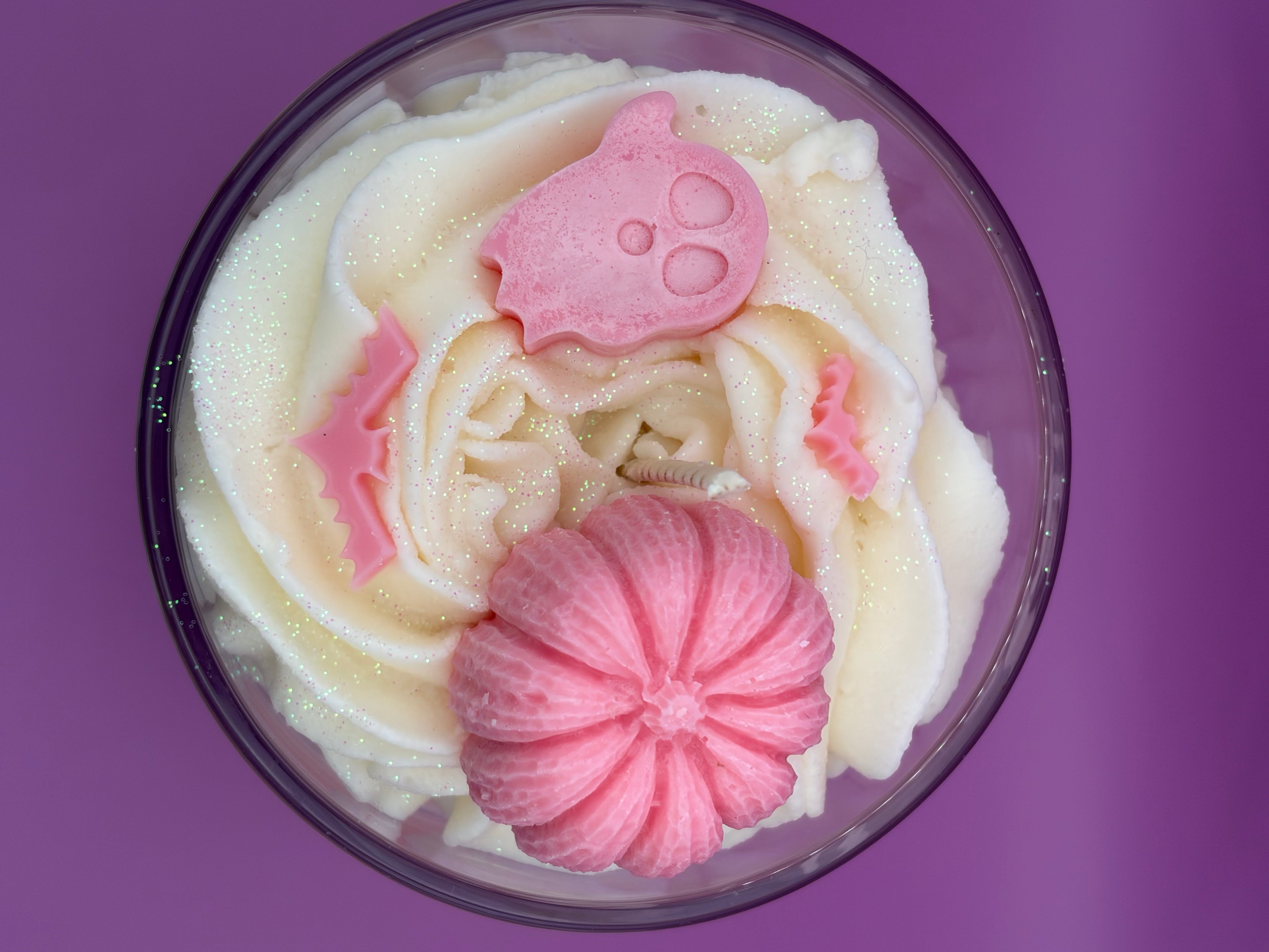 A top-down view of a clear glass candle filled with white whipped frosting, decorated with pink shapes including a ghost and pumpkin, set against a solid purple background.