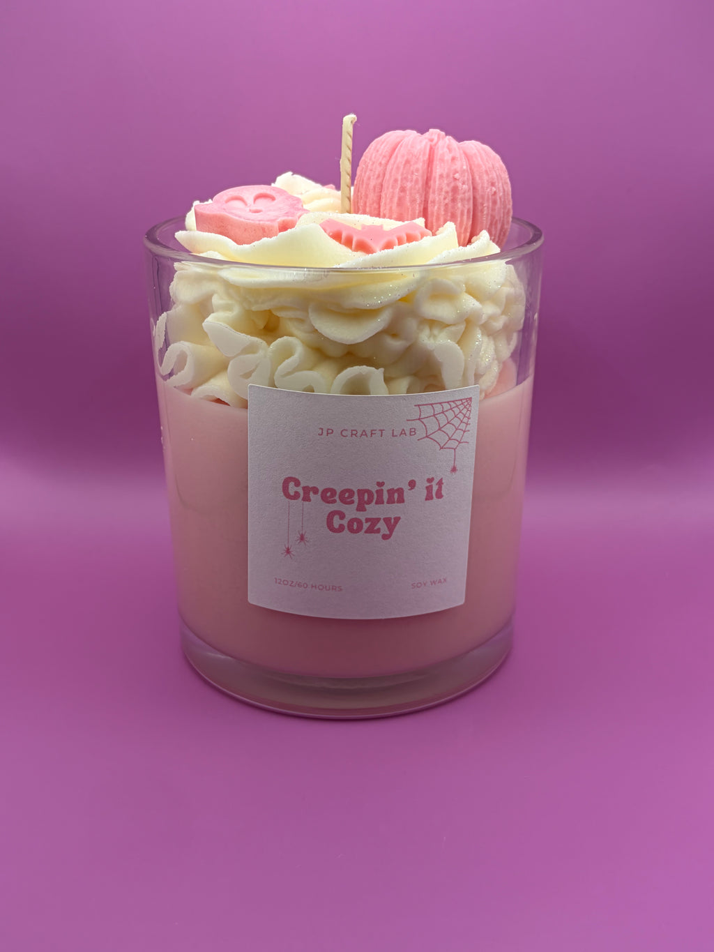 A pink candle in a clear glass jar with a white label reading 'Creepin' it Cozy' and 'JP CRAFT LAB'. The top of the candle is decorated with cream-colored ruffled wax, a pink macaron wax piece, and a small pink pumpkin wax ornament, all set against a solid purple background.