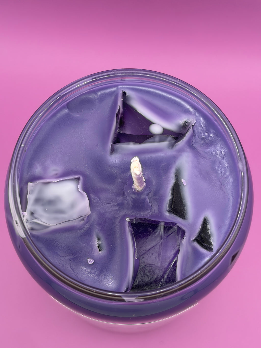 A round purple candle in a clear glass container, photographed from above against a solid pink background. The top of the candle has a textured, cracked ice cube surface with a single white wick visible in the center.