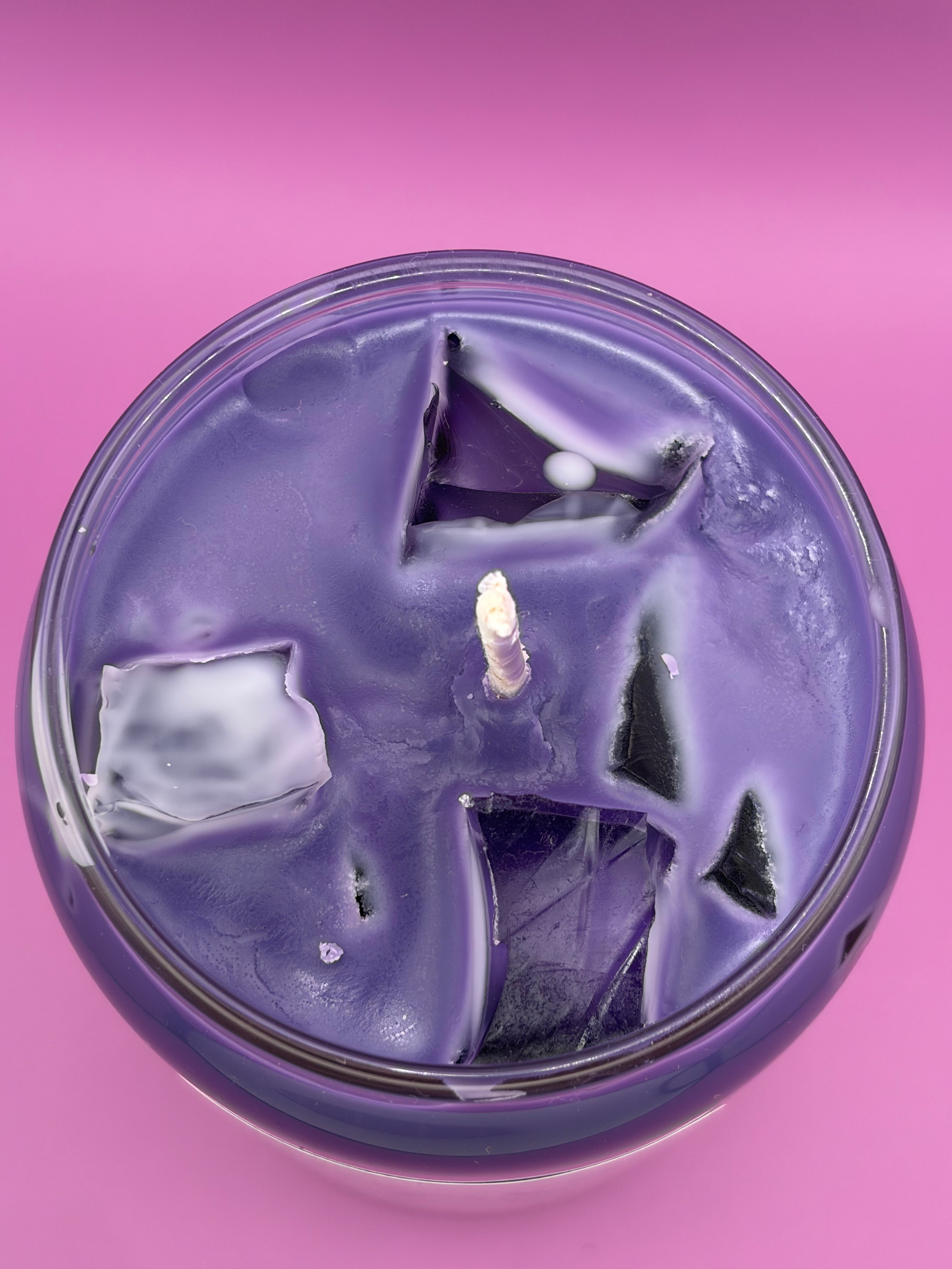A round purple candle in a clear glass container, photographed from above against a solid pink background. The top of the candle has a textured, cracked ice cube surface with a single white wick visible in the center.