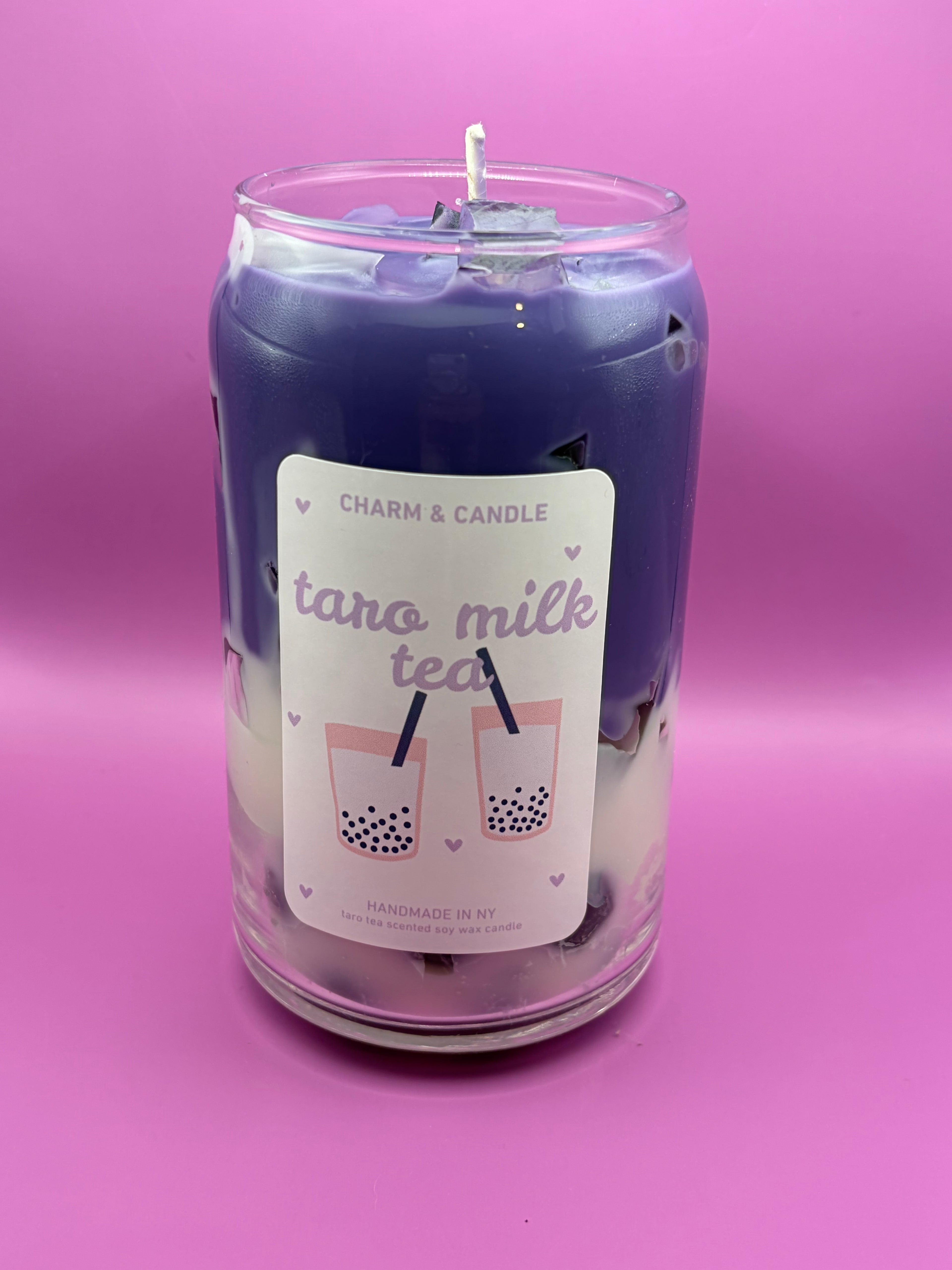 A glass jar candle with a purple wax top layer and a white wax bottom layer, featuring a white label that reads 'taro milk tea' with illustrations of two boba tea cups. The candle is set against a solid pink background.