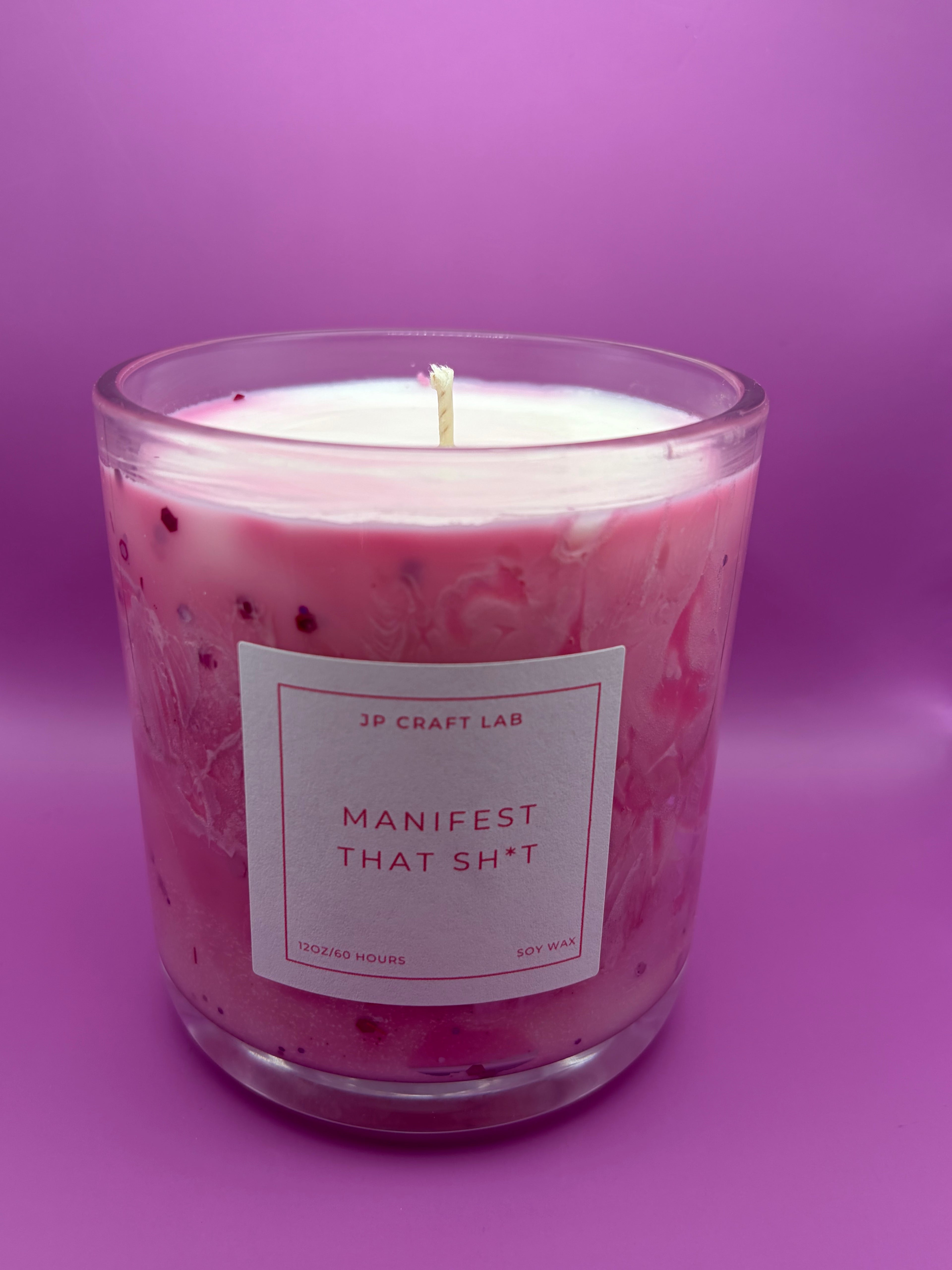 A pink candle in a clear glass jar with a white label reading 'JP CRAFT LAB MANIFEST THAT SH*T' on a solid purple background.
