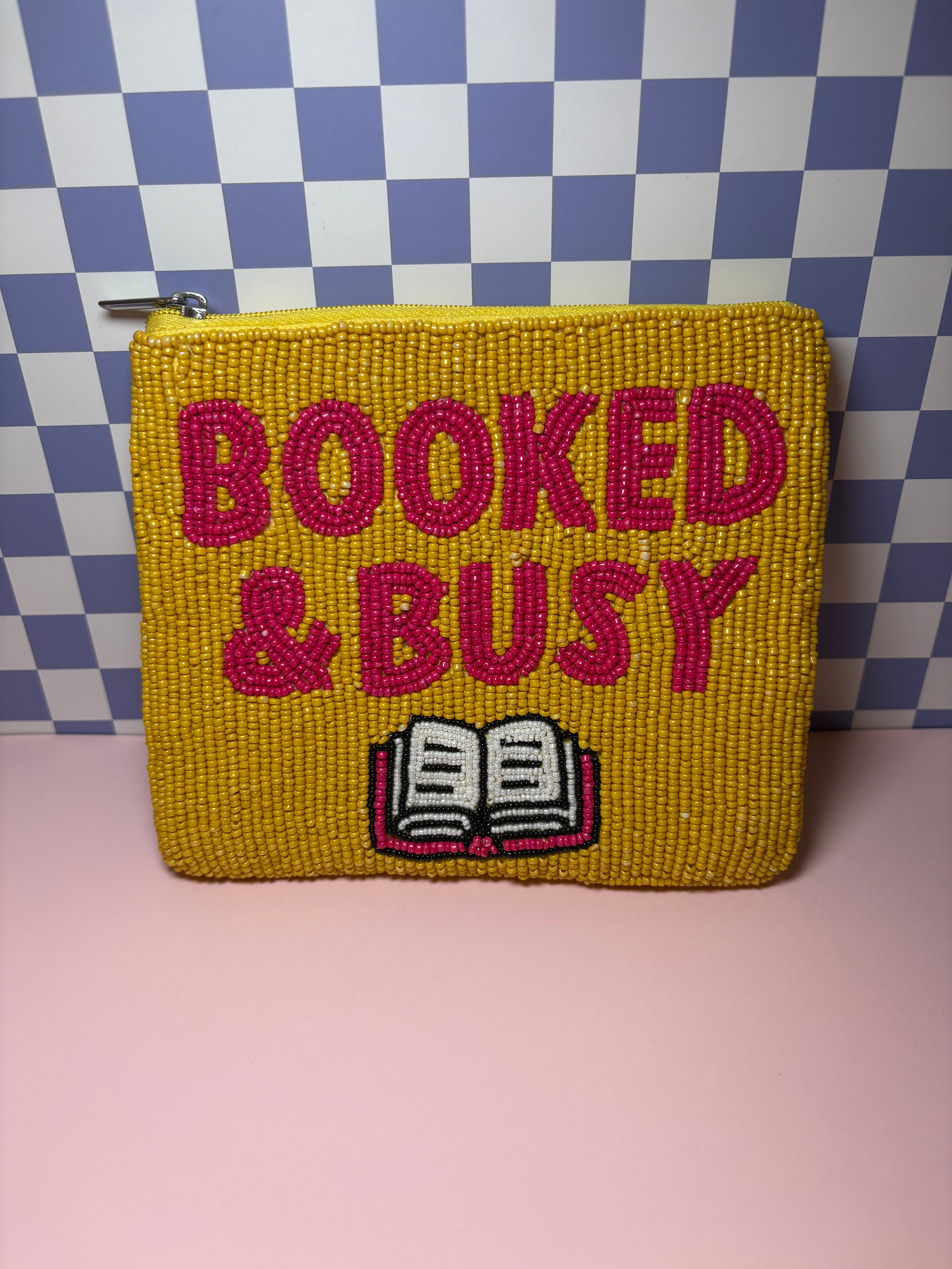 A yellow beaded zippered pouch with pink text reading 'BOOKED & BUSY' and a small book icon, set against a blue-and-white checkered background and pink surface.
