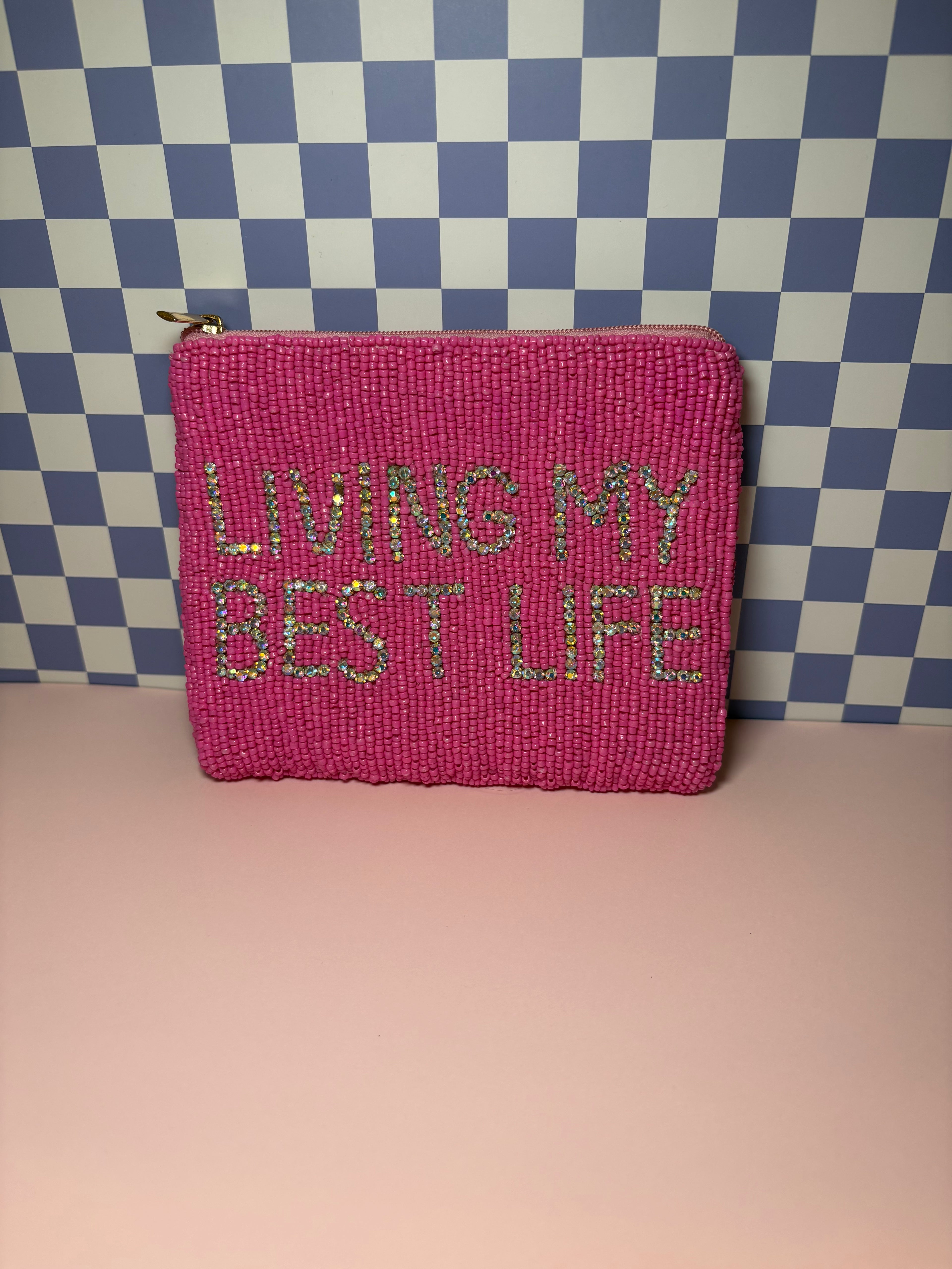 A bright pink beaded zippered pouch with the phrase 'LIVING MY BEST LIFE' in glittery silver lettering, set against a blue and white checkered background and a light pink surface.