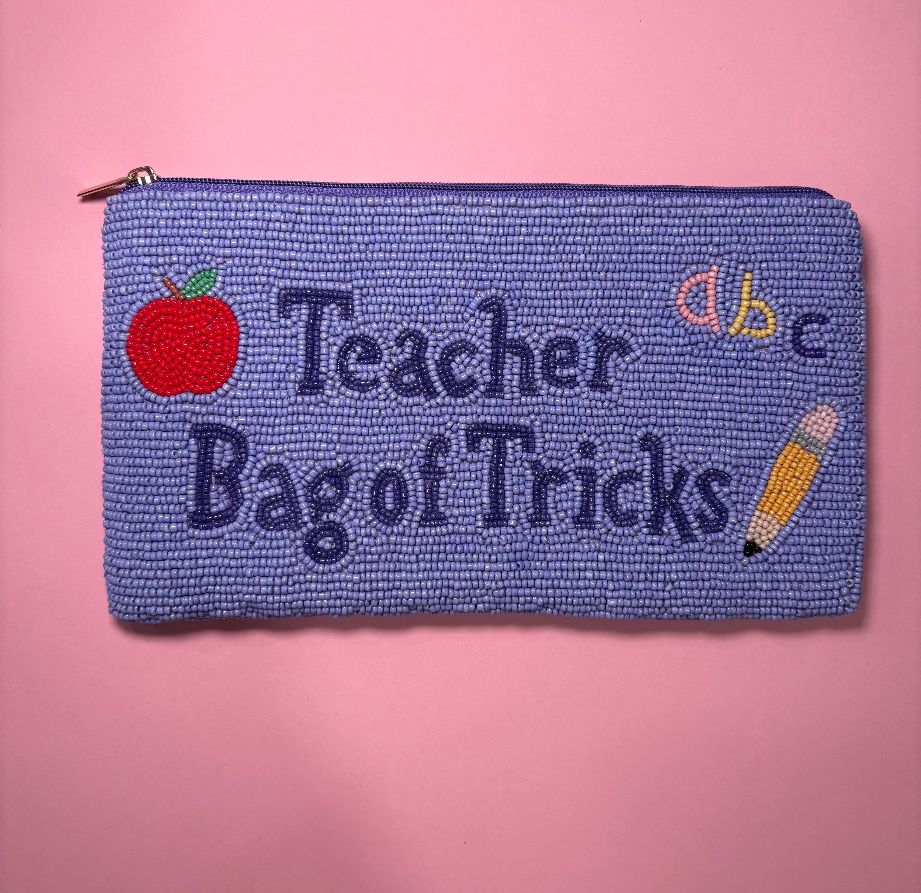 A purple beaded zipper pouch with 'Teacher Bag of Tricks' embroidered in purple, featuring a red apple, a yellow pencil, and colorful letters on a pink background.