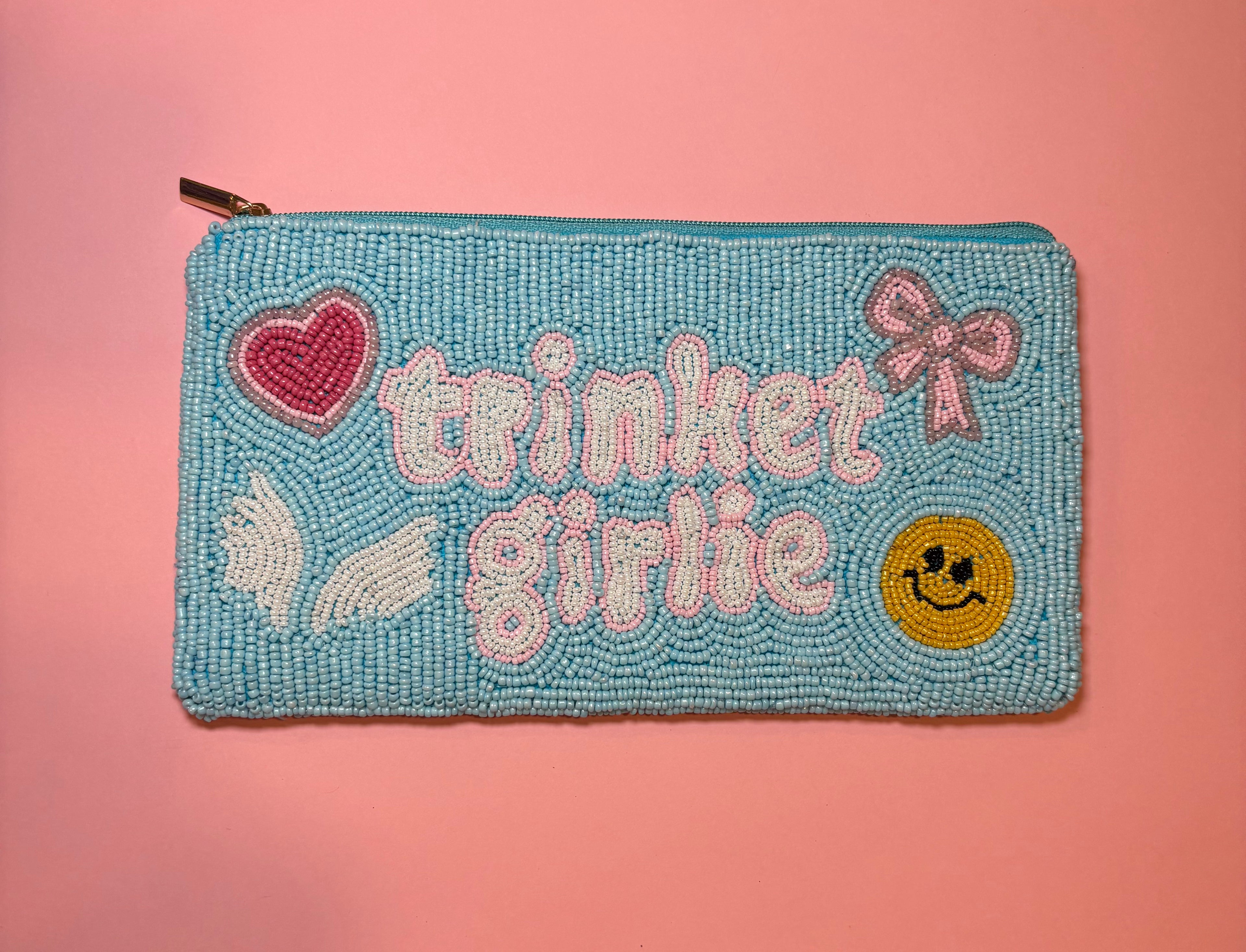A rectangular beaded zipper pouch in light blue with the words 'trinket girlie' in pink and white, decorated with a pink heart, white wings, a pink bow, and a yellow smiley face, shown against a solid pink background.