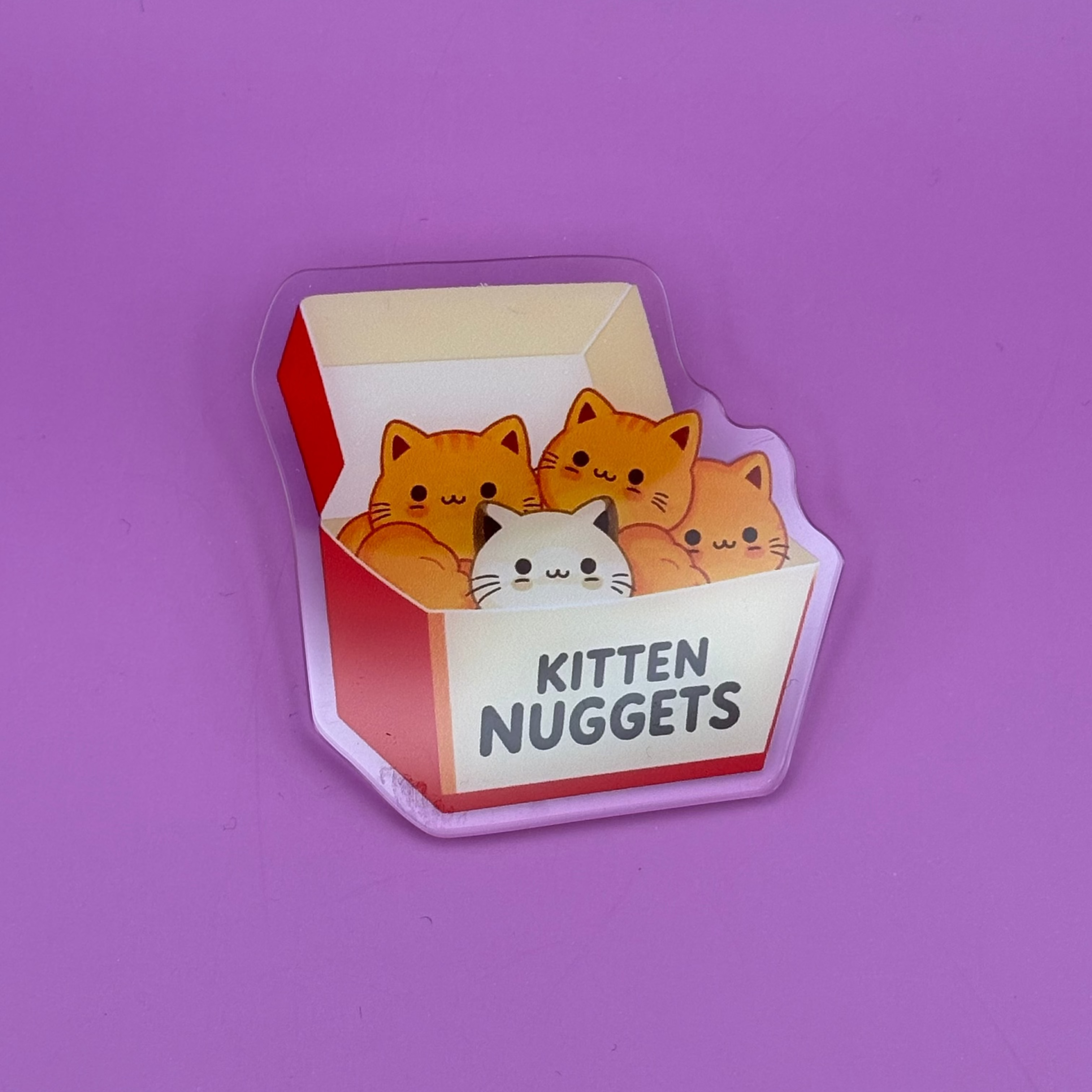 A novelty sticker or magnet shaped like a cigarette pack, featuring cartoon orange and white kittens peeking out with the text 'KITTEN NUGGETS' on a purple background.