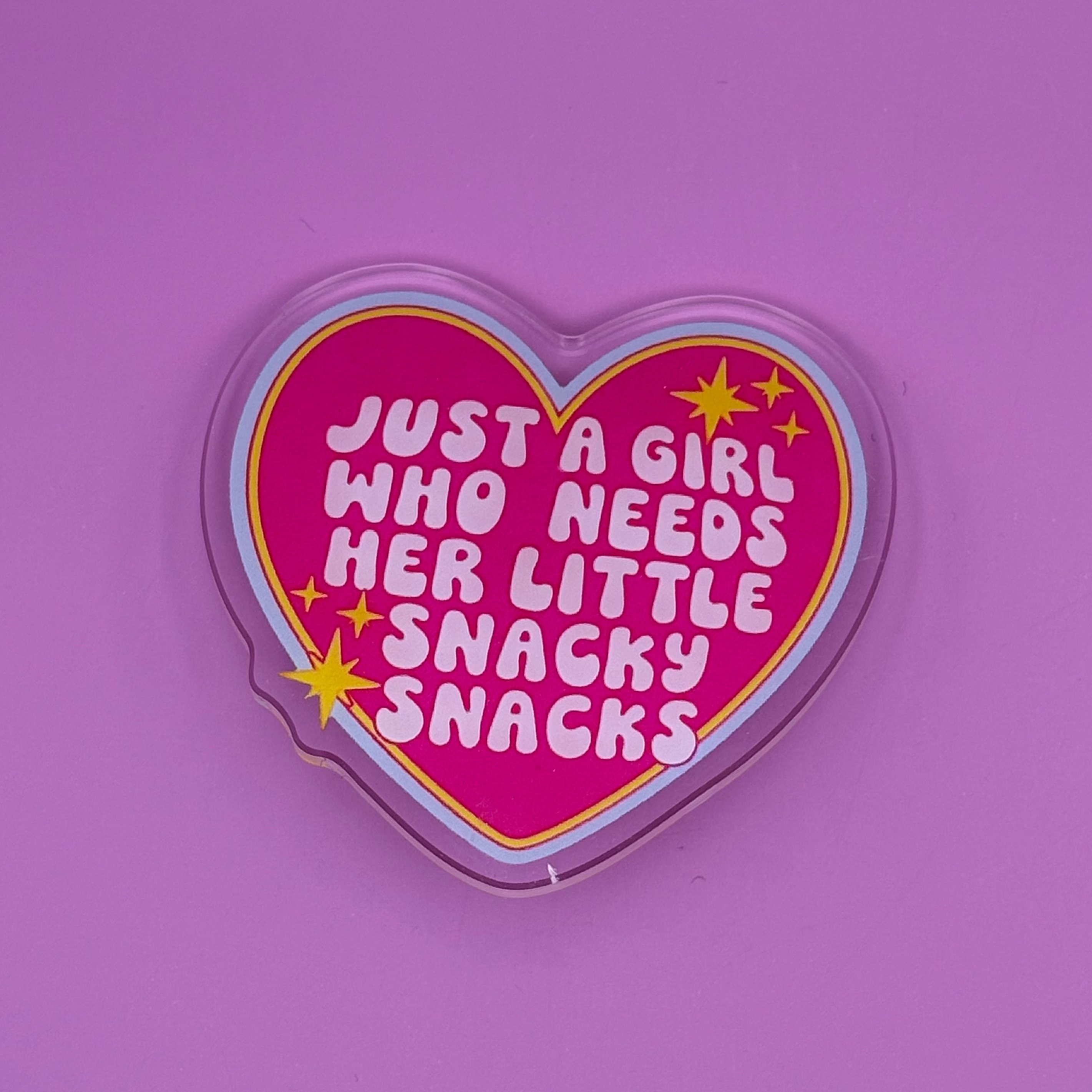A heart-shaped clear plastic sticker or decal with a hot pink background and white text that reads 'JUST A GIRL WHO NEEDS HER LITTLE SNACKY SNACKS' with yellow stars, shown on a solid purple background.