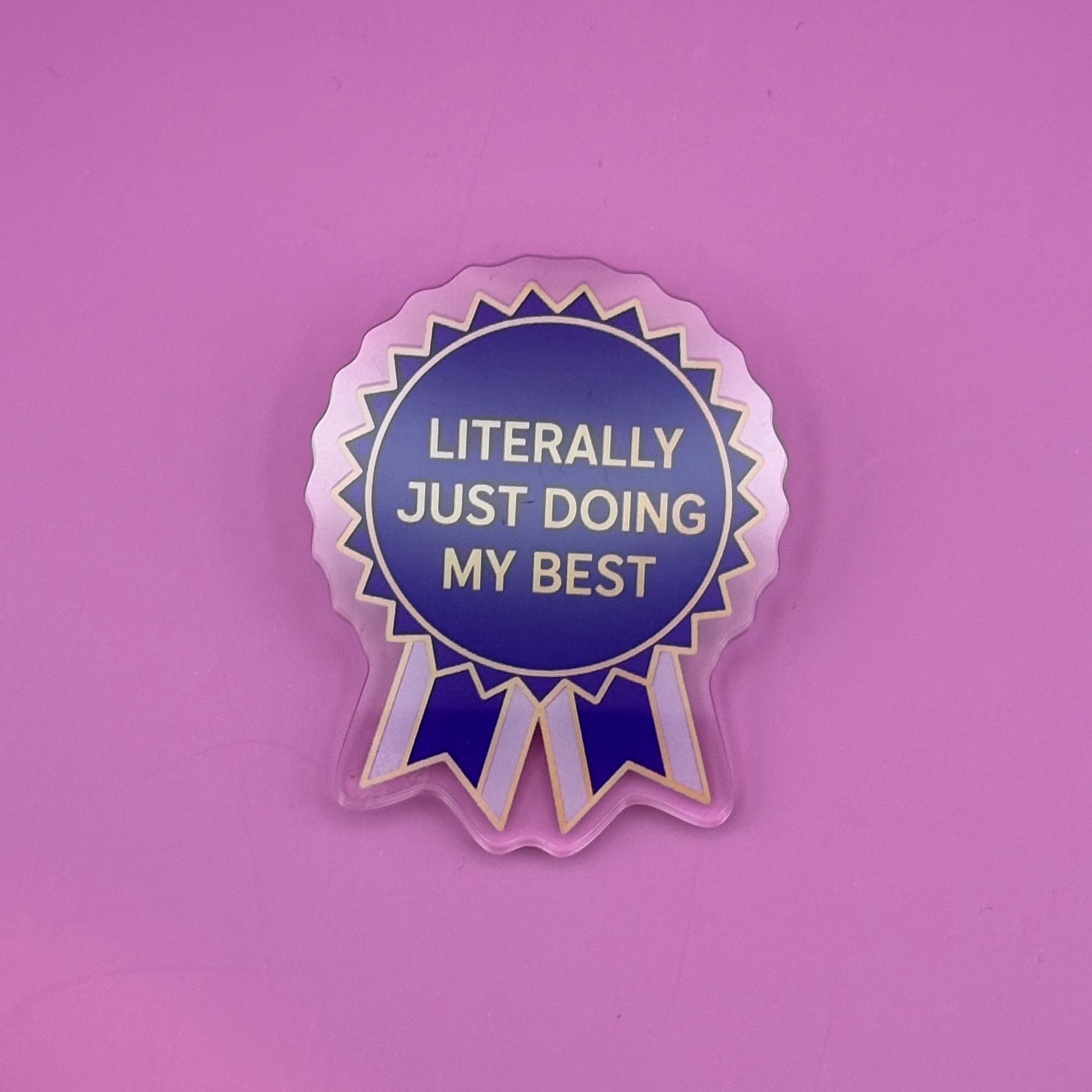 A die-cut sticker shaped like a blue and white ribbon award badge with gold text reading 'LITERALLY JUST DOING MY BEST', shown on a solid pink background.